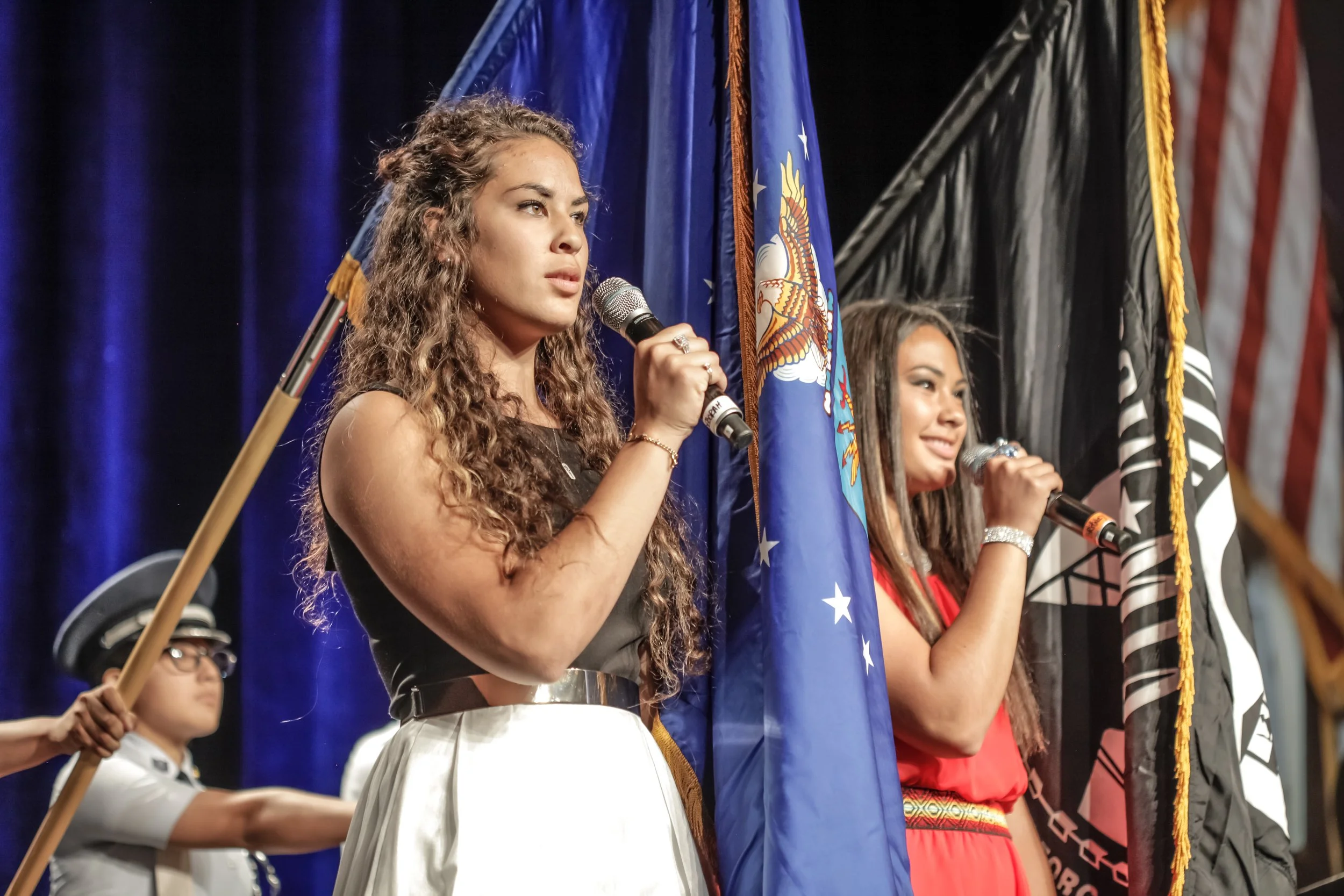 Two young women hold flags and microphones during a ceremony or event, with a woman in the background holding a flag.