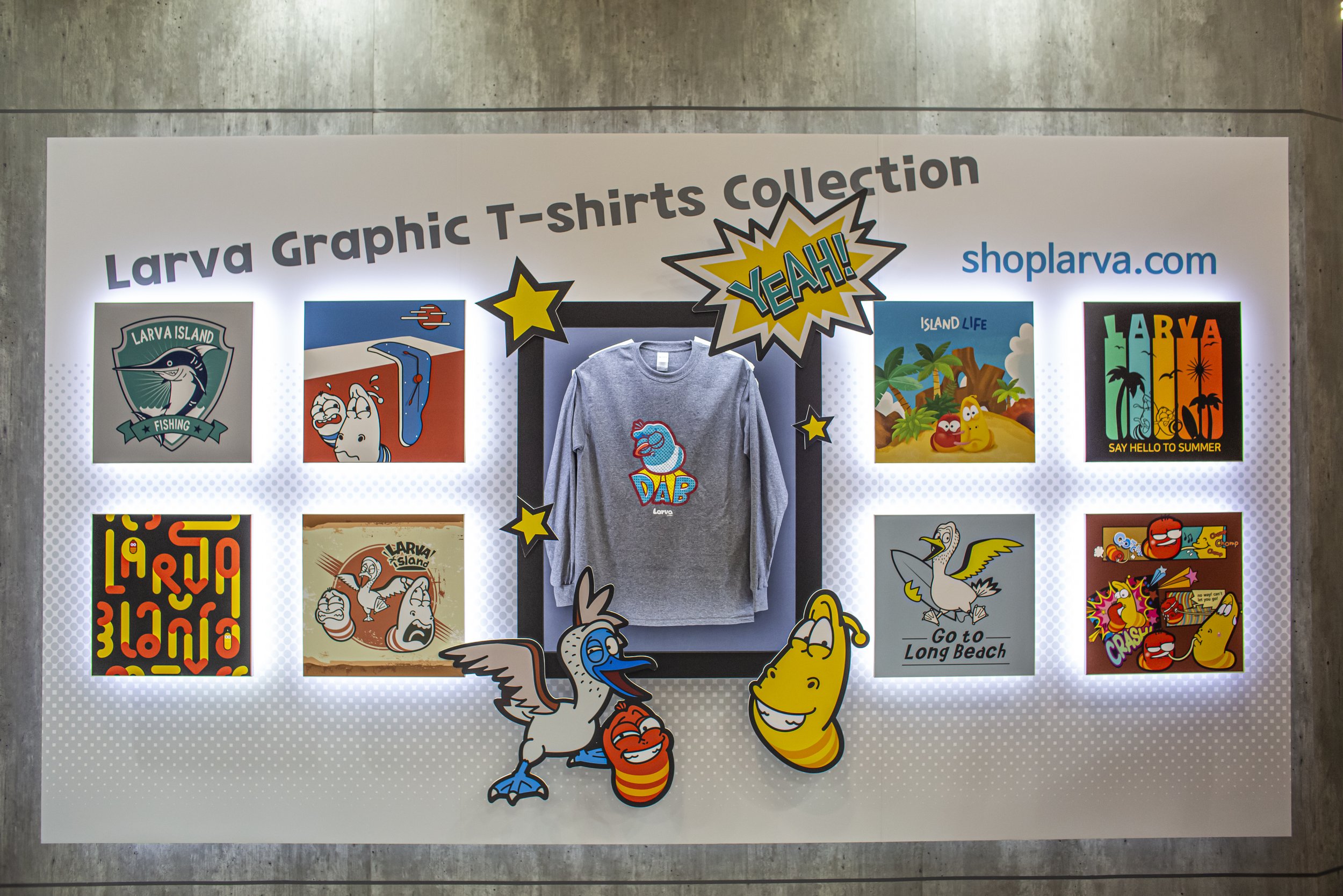 Display of Larva graphic T-shirts collection with designs featuring cartoon characters and tropical themes, as well as a central gray T-shirt with a cartoon dabbing character. Bright decals and illustrations highlight the collection. Line Friends.