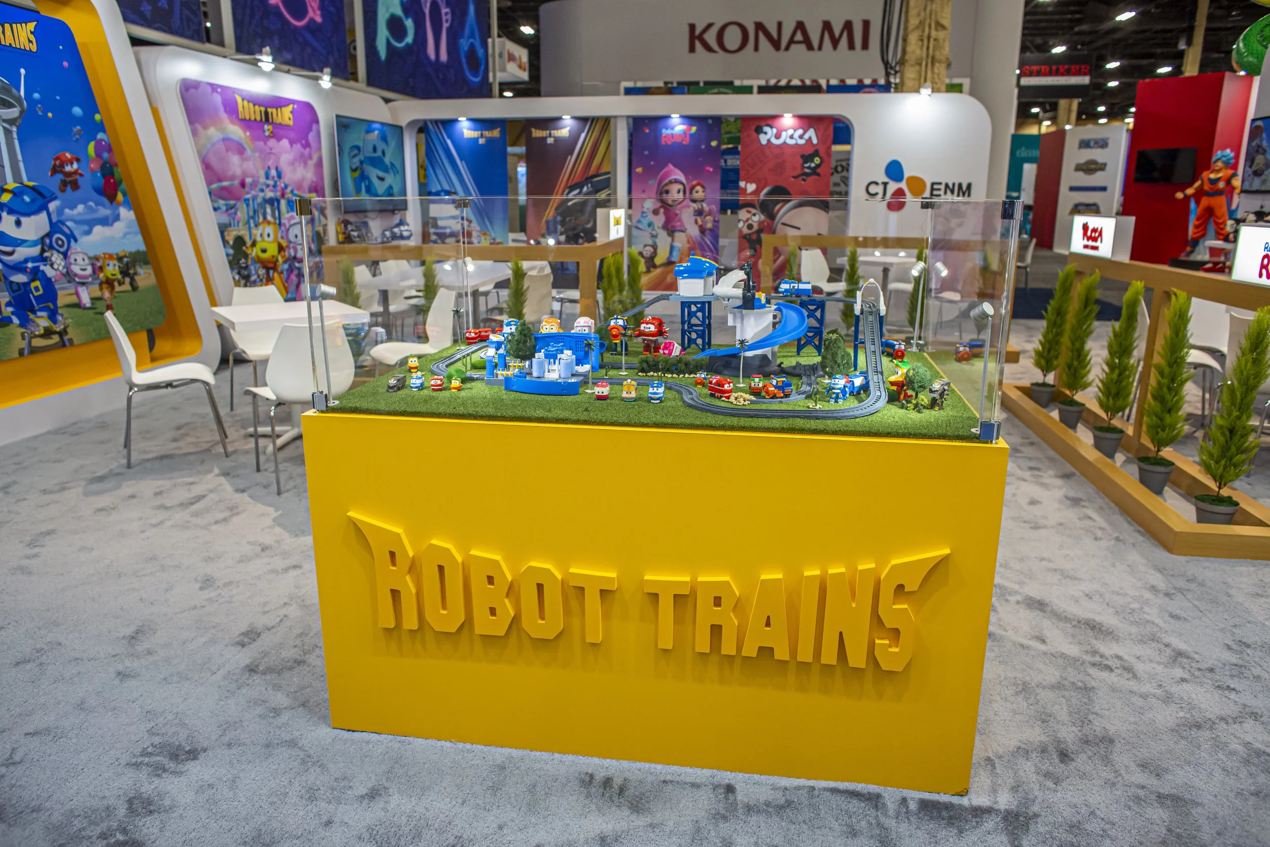 A scale model of a toy train park display inside a convention booth with a yellow sign that reads "BUILDING TOYS". The display includes small toy trains, trees, and tracks. In the background, there are posters and banners for various toy brands inclu