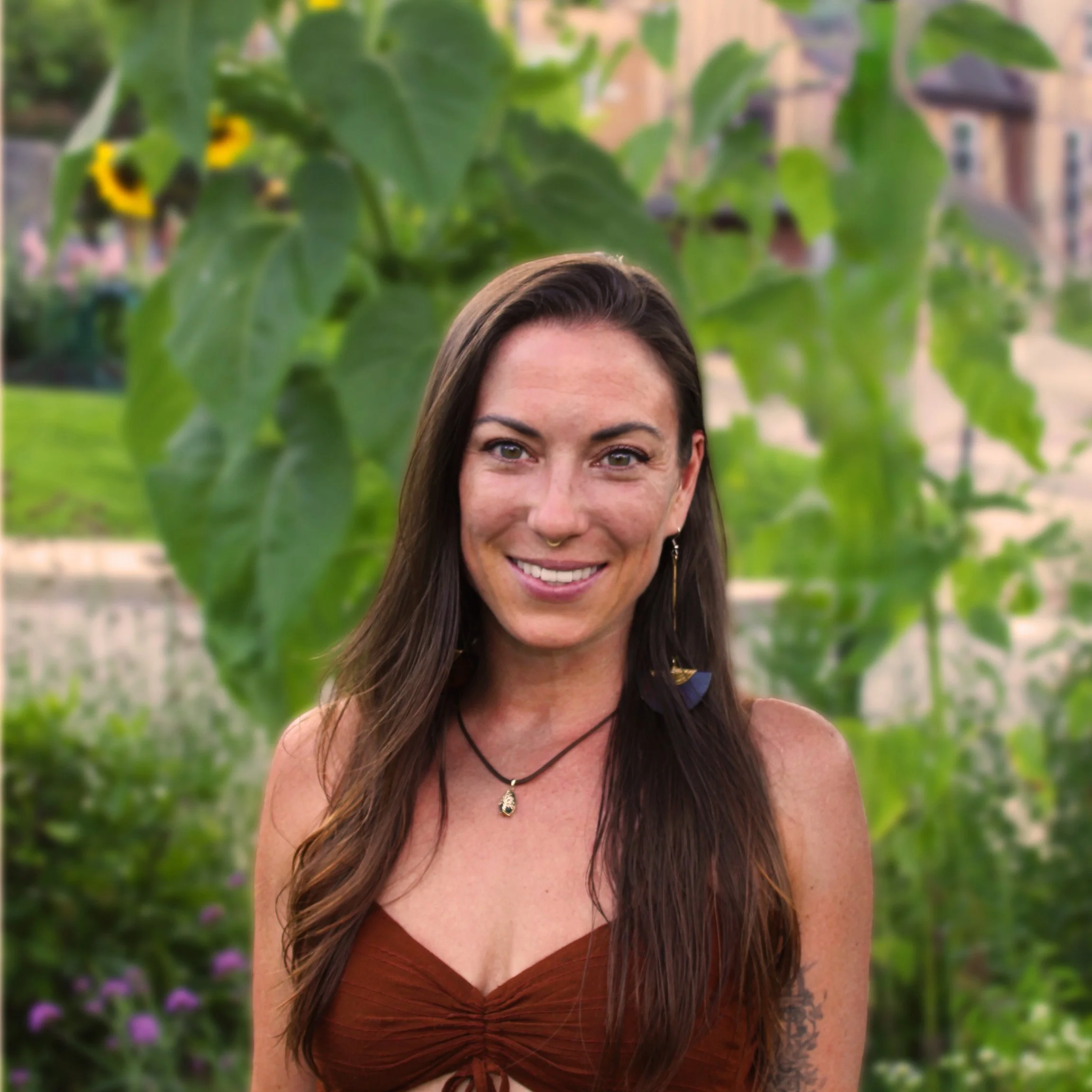 A woman with long brown hair smiling outdoors, standing in front of green leaves and colorful flowers with a blurred background of a city park or garden.
