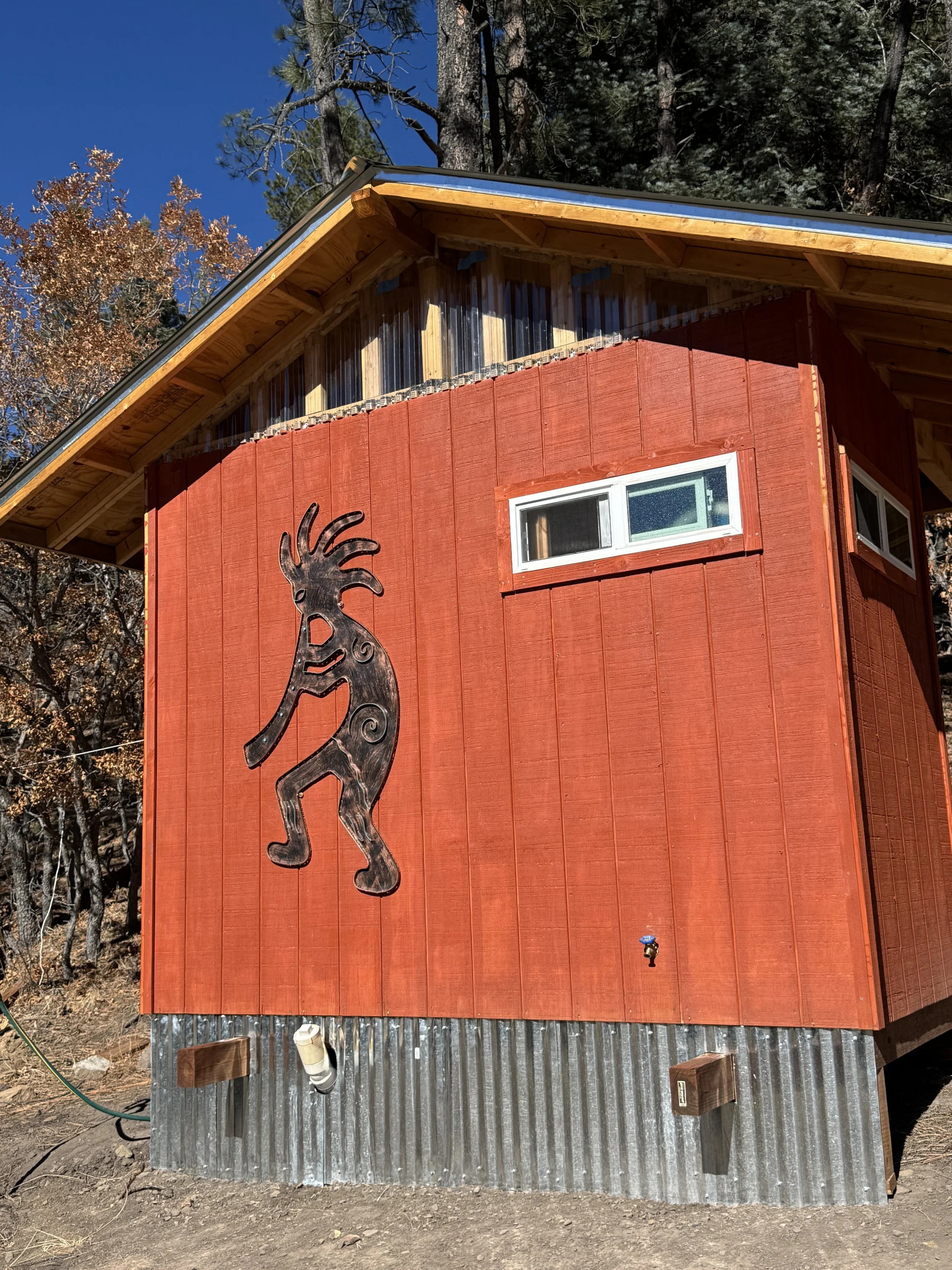 A small shower area with corrugated metal walls, a pebble-textured shower floor, a white strip shower curtain on a rod, a wall-mounted white heater, and a striped bath mat outside the shower.