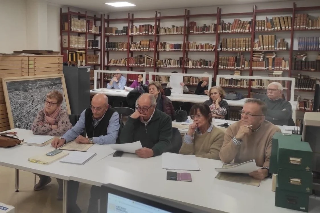 Grupo de personas adultas en una sala de reuniones con estanterías llenas de libros en el fondo, algunos toman notas y otros escuchan atentamente.