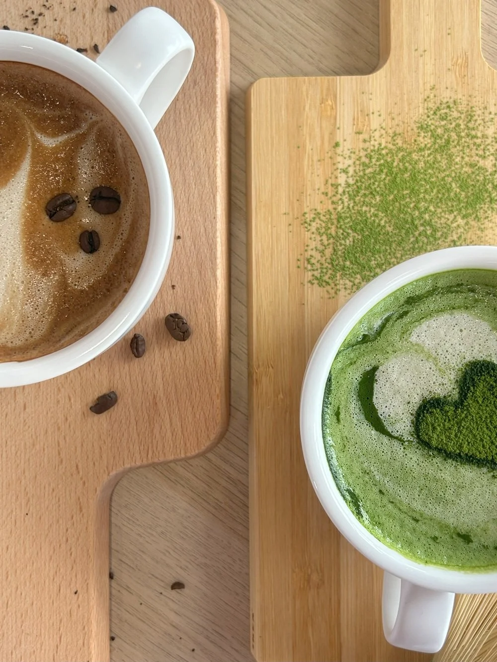 Two cups of coffee, one with brown foam and coffee beans on top, and the other with green matcha tea, placed on wooden boards.