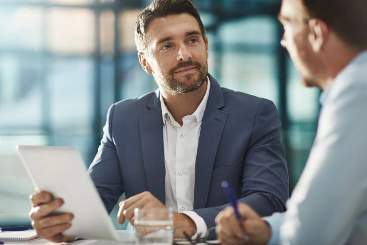 Two businessmen in a professional setting, one with a tablet and the other taking notes, engaged in a conversation.
