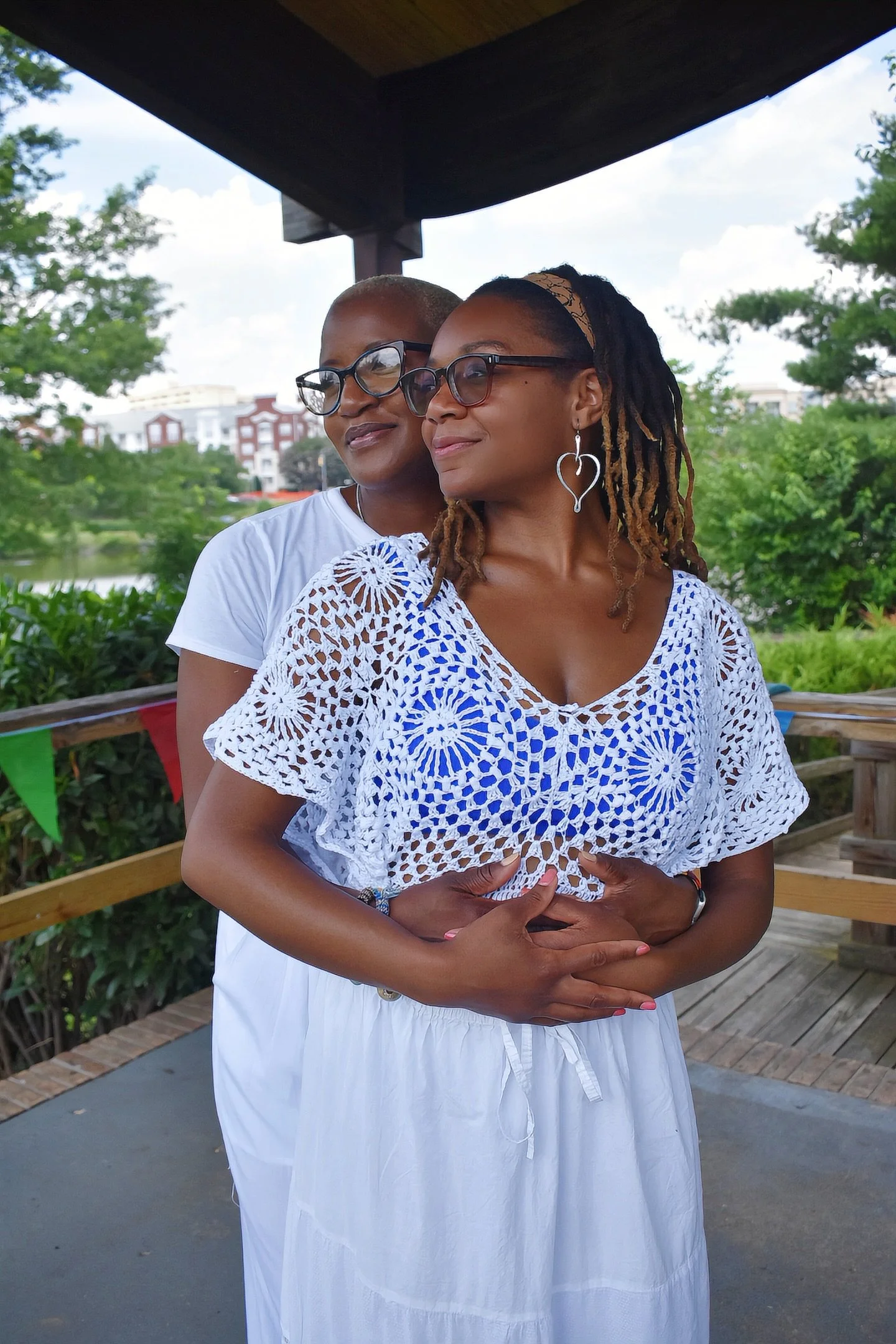 Two women standing outdoors, one behind the other, with the woman in the front holding her hands over her abdomen. Both are wearing glasses and white clothing, with greenery and buildings in the background.