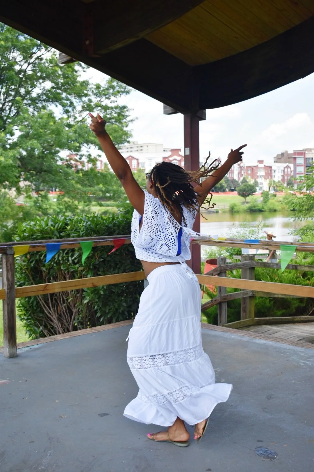 A woman with dreadlocks dancing outdoors on a pavilion stage, wearing a white skirt and crochet top, with trees, water, and buildings in the background.