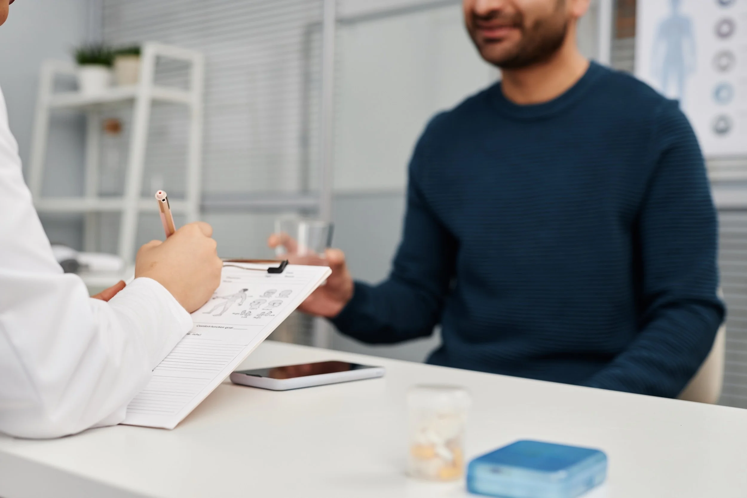 A man in a blue shirt receiving a medical consultation from a healthcare professional holding a clipboard and pen in a clinical setting.