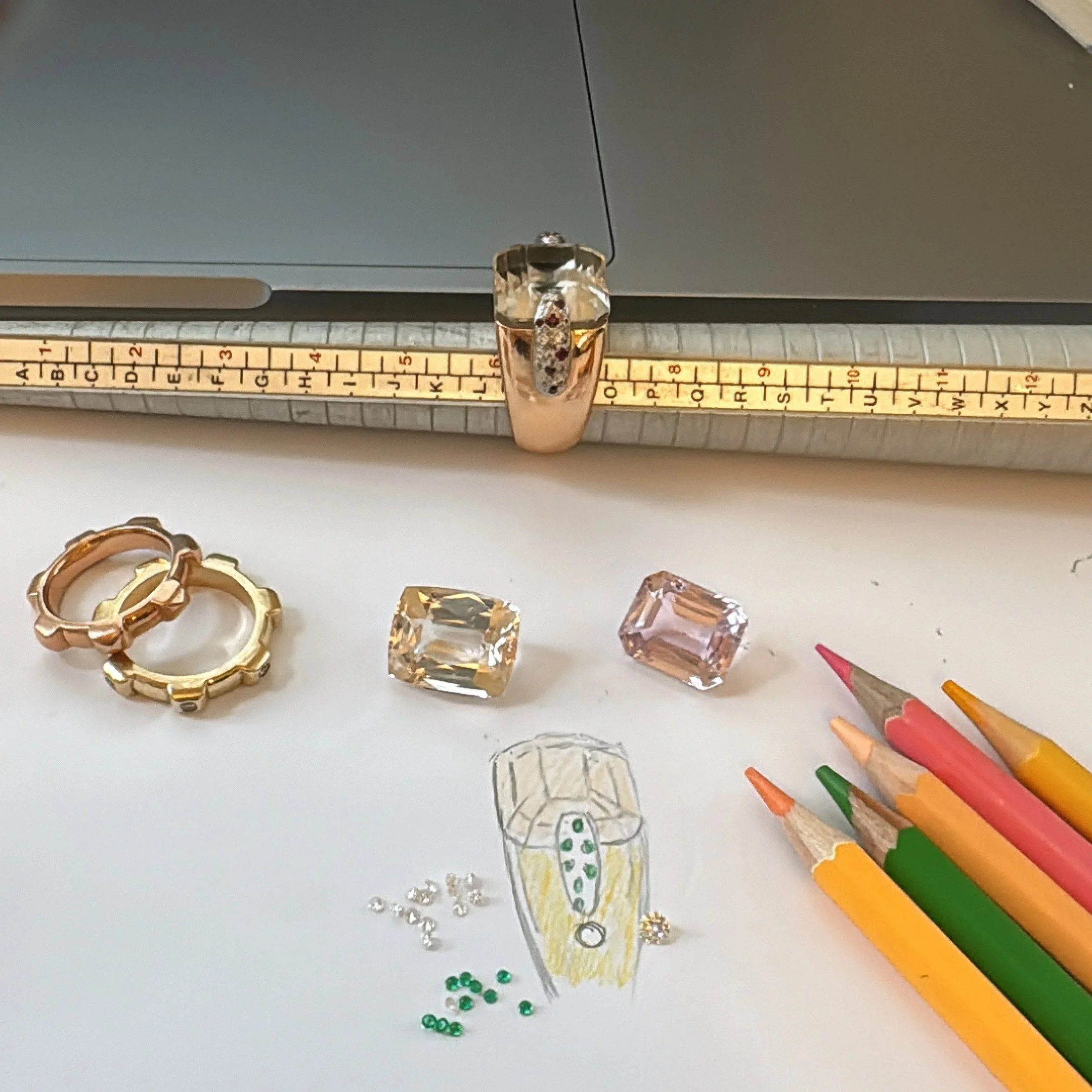 Bespoke jewellery design process with loose gemstones, coloured pencils, gold rings and a hand-drawn sketch on the workbench