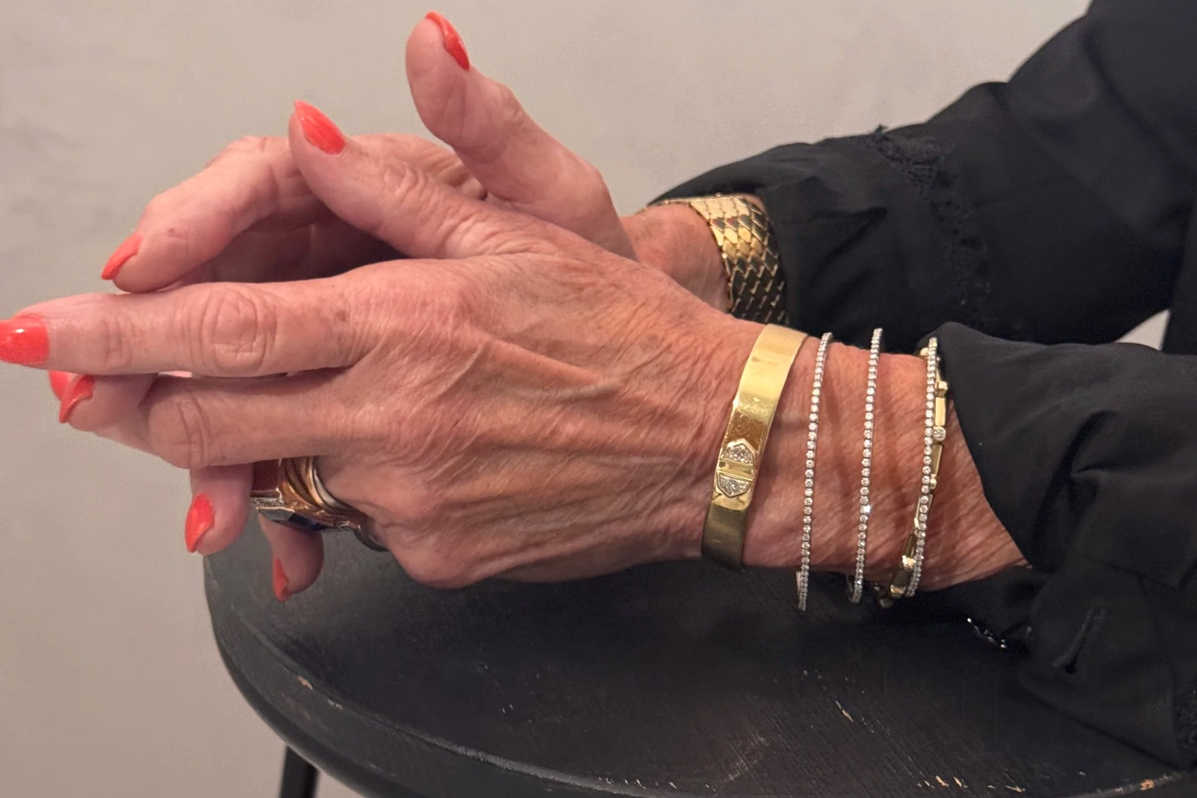 Close-up of a woman wearing layered gold necklaces, large rings with a clear rectangular gemstone, and a black strap top, inside a modern room with reflective wall panels and a window in the background.