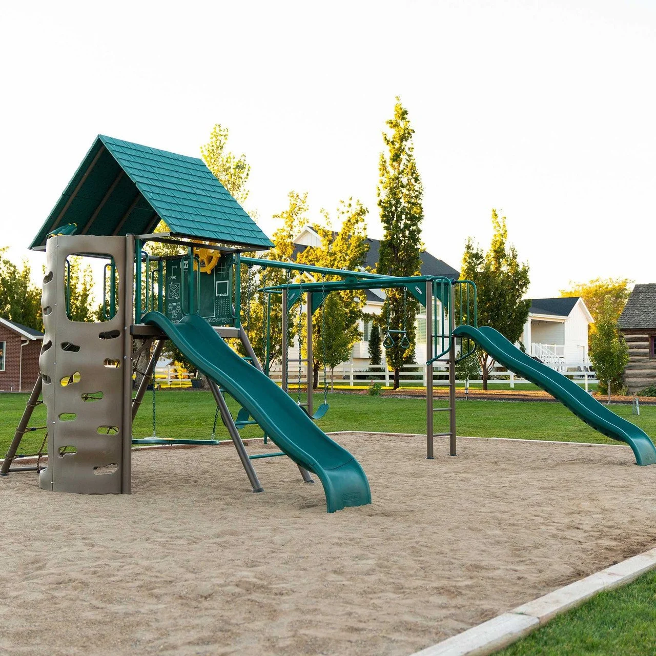 Playground with slides, swings, and a small playhouse with a blue shingled roof on sand surface, surrounded by grass and trees.
