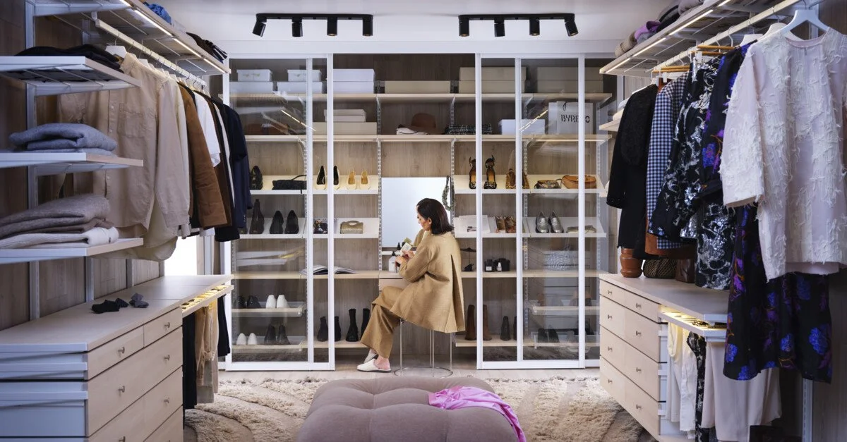 A woman sitting on a stool in a walk-in closet surrounded by shelves of shoes, clothing, and accessories.