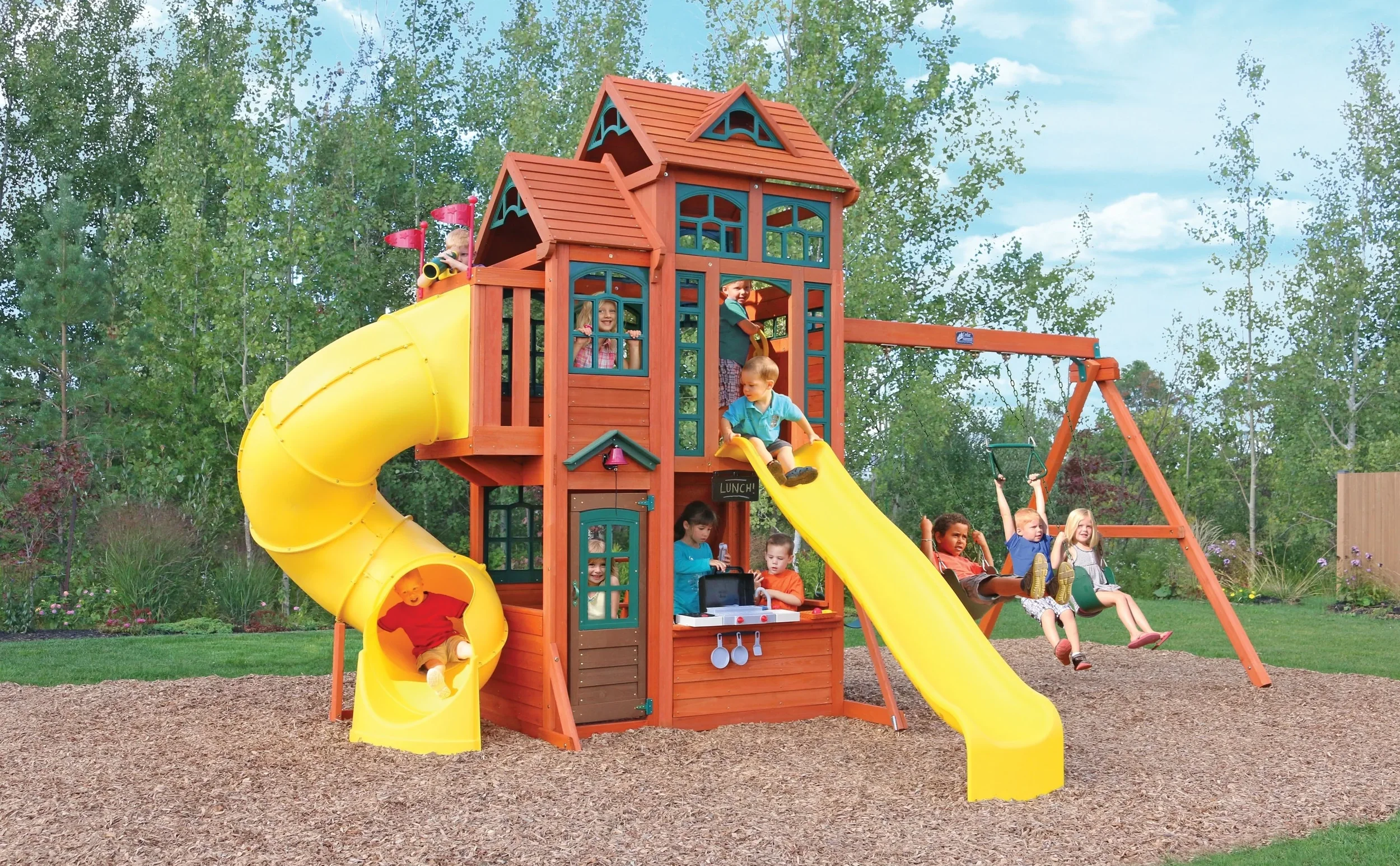 Children playing on a wooden playground with slides, swings, and a playhouse in a grassy yard.