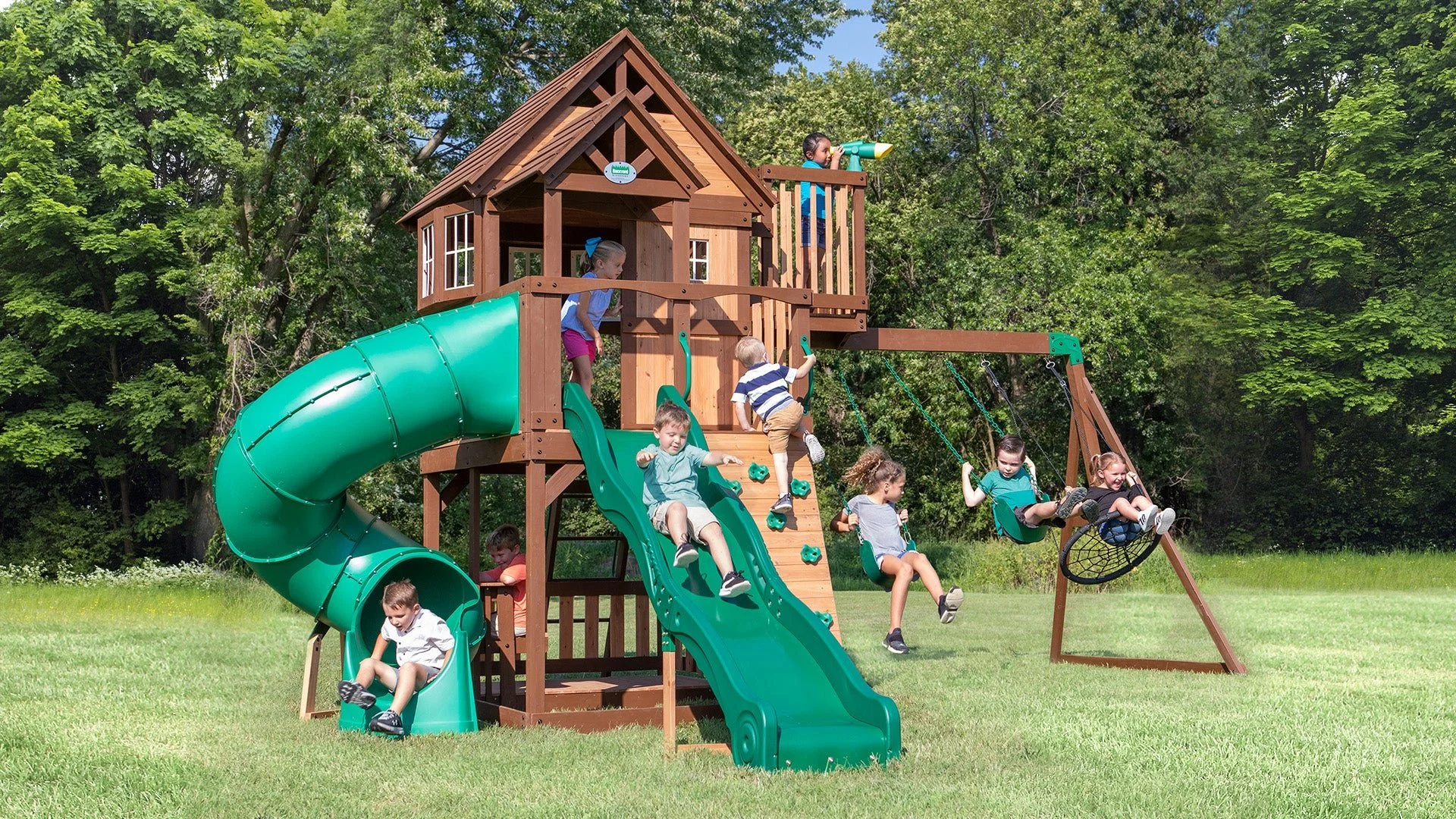Children playing on a large wooden playground set with slides, swings, and climbing wall in an outdoor park with trees.