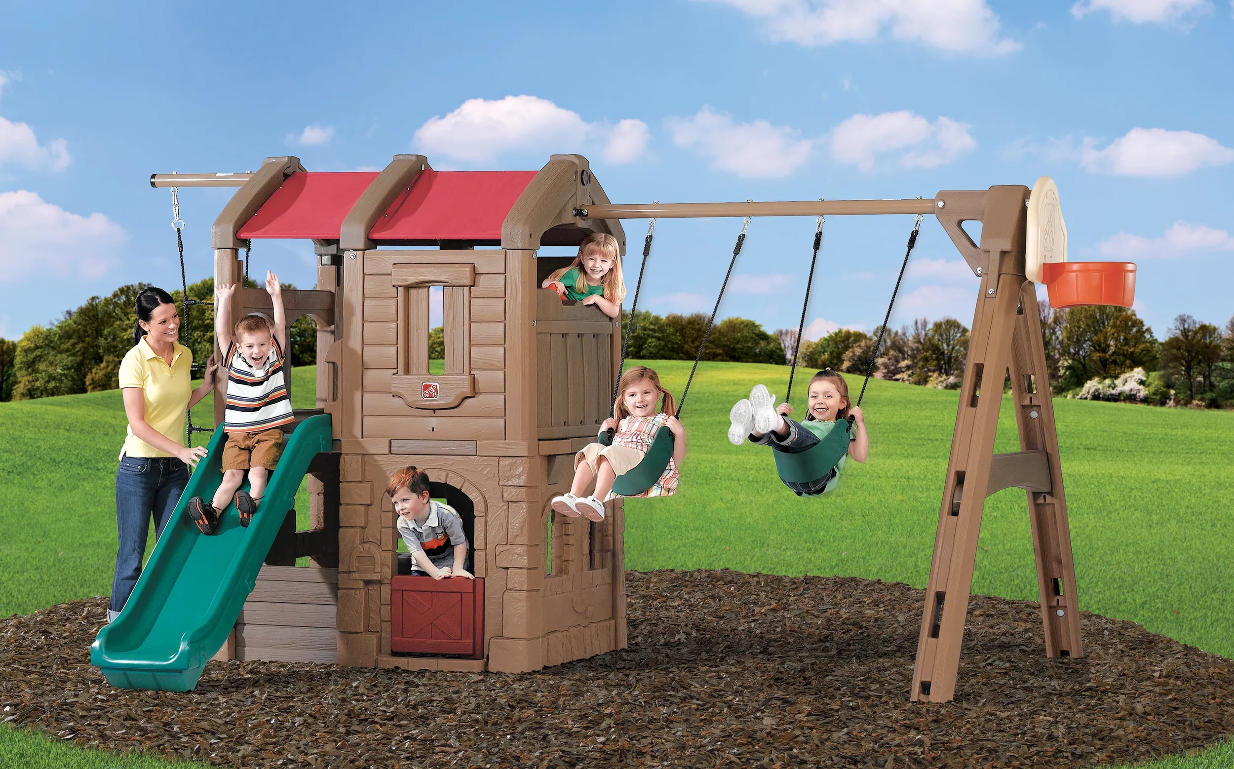 Children playing on a large plastic playground set with a slide, swings, and a playhouse in a grassy outdoor area under a blue sky.