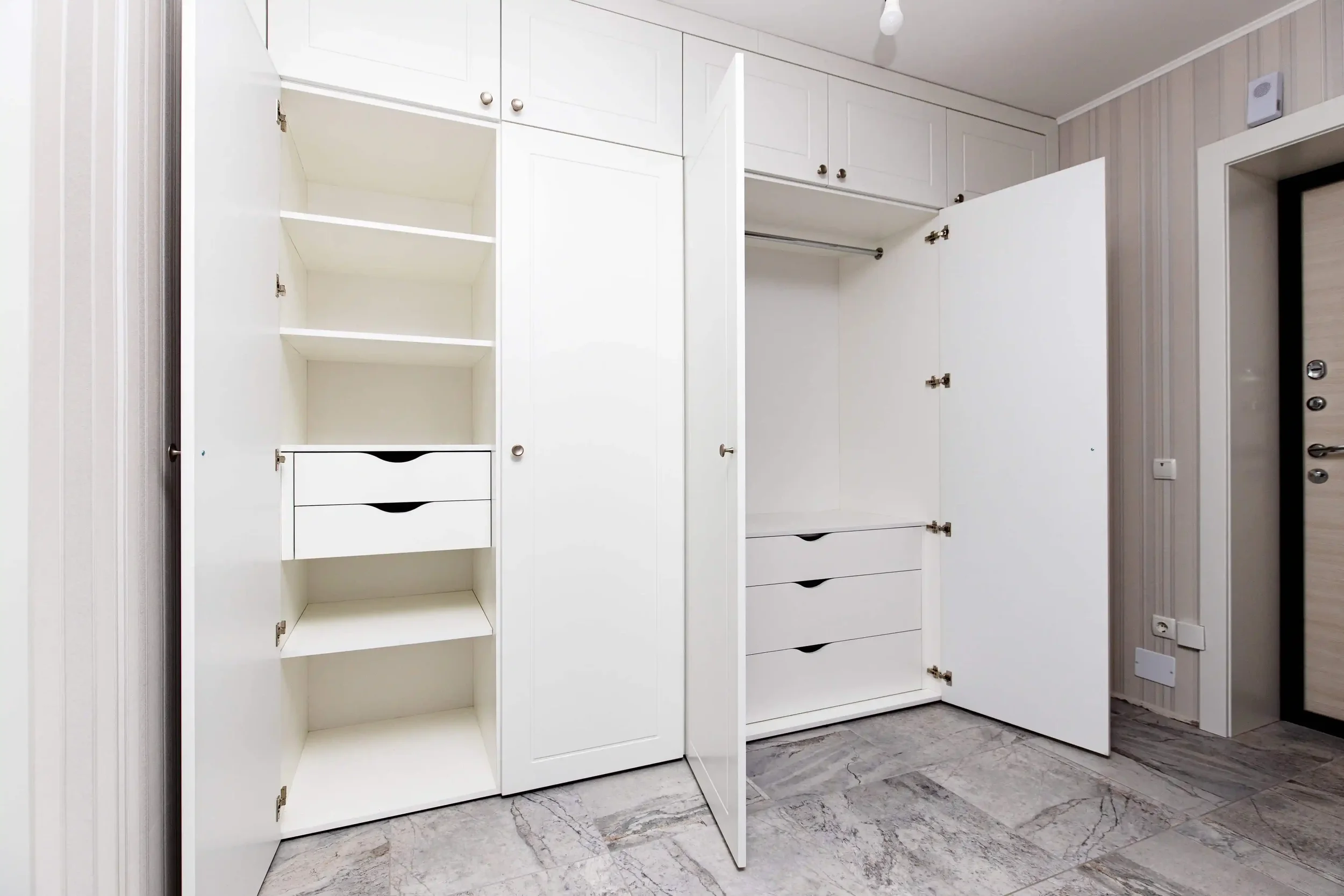 Empty white built-in closet with shelves and drawers in a room with beige striped wallpaper and gray tile floor.