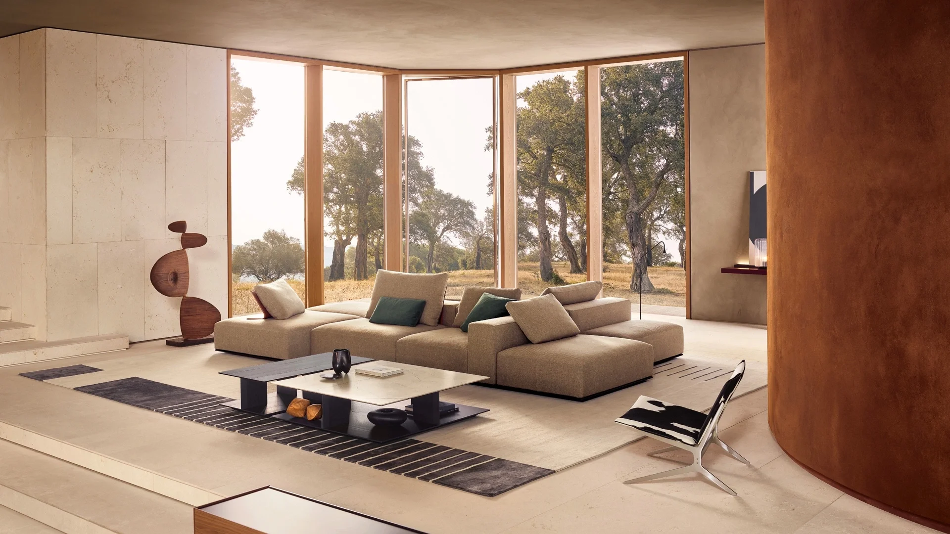 Modern living room with large windows overlooking a natural landscape, beige sofa with pillows, black and white chair, coffee table, decorative sculpture, and minimal decor.