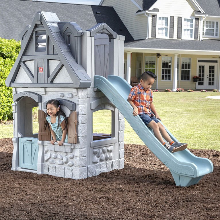 Two children playing on a plastic playhouse with a slide in a backyard, with a house and green lawn in the background.