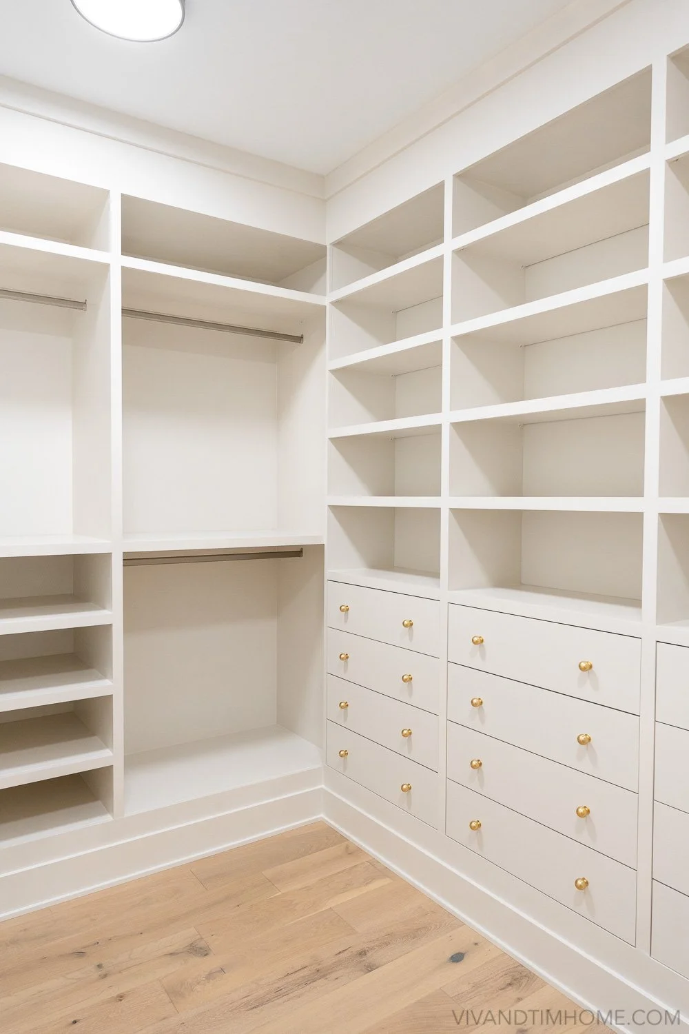 Empty white walk-in closet with built-in shelves, drawers with gold knobs, and hanging rods on a wooden floor.