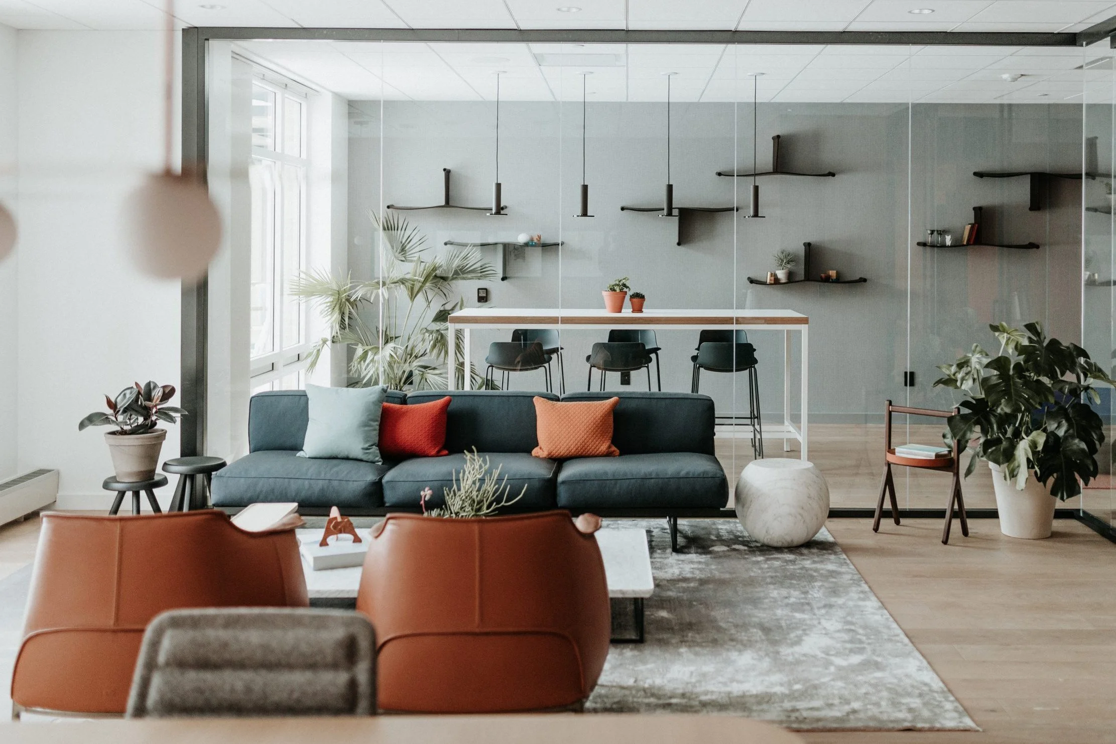 Modern office lounge area with a dark sofa, orange and light blue pillows, surrounding plants, a coffee table, and a glass partition separating a workspace with bar stools and a counter.