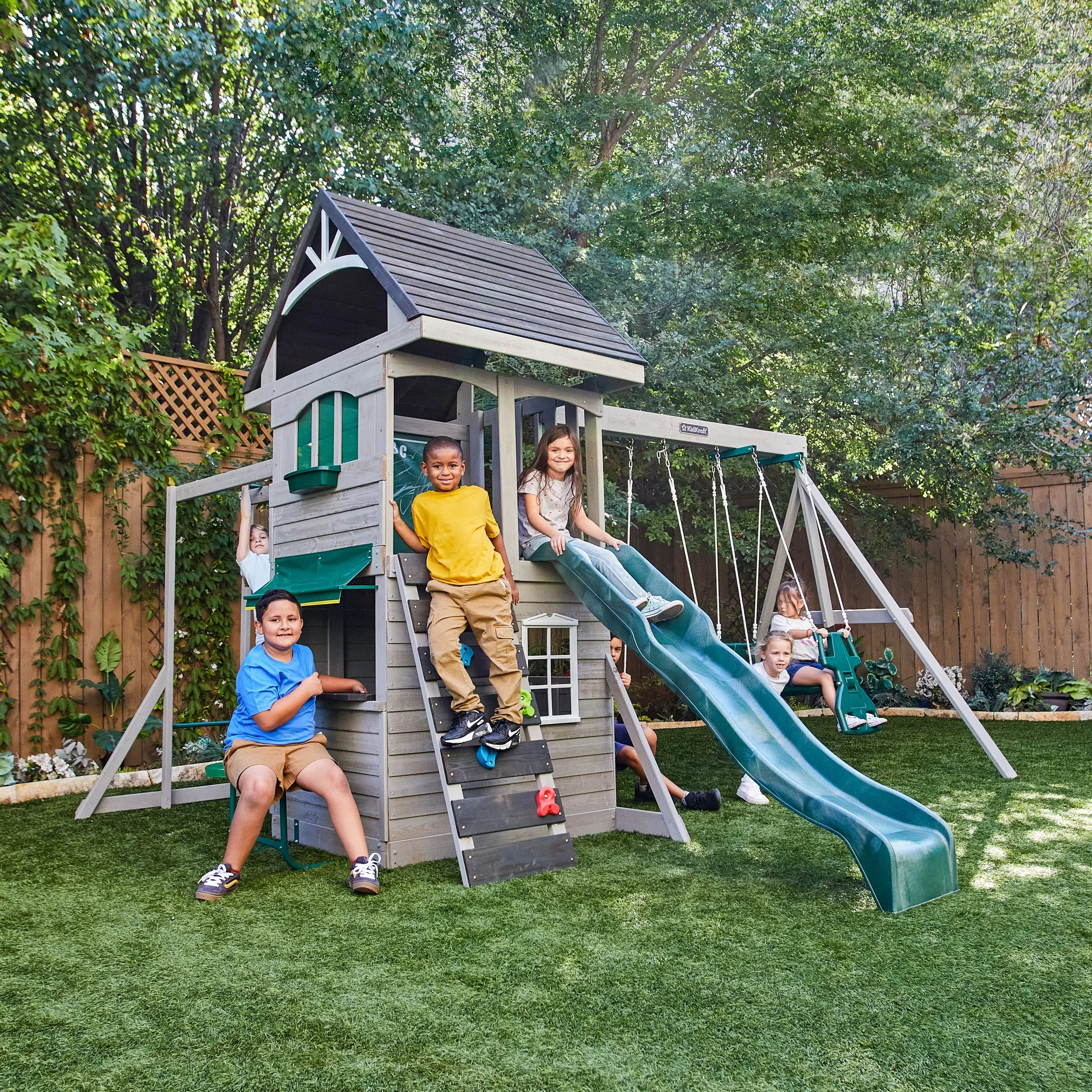 Children playing on a wooden backyard playset with slides and swings, surrounded by green trees and a fenced yard.