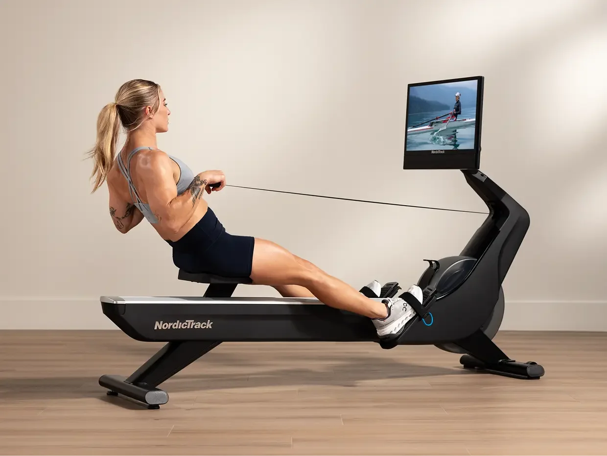 A woman exercising on a NordicTrack rowing machine in a room with wooden floor and neutral-colored wall.