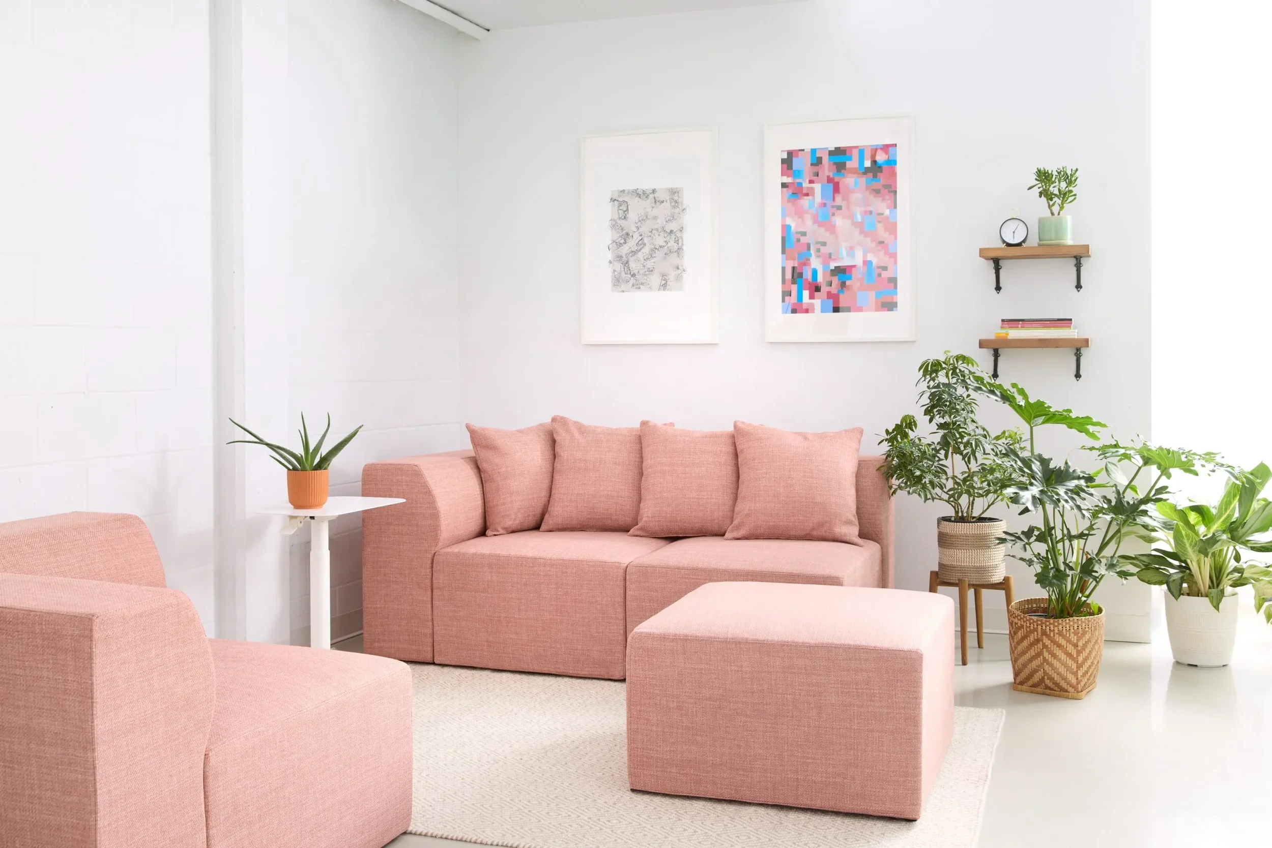 A minimalist living room with a pink sofa, matching pink armchair, and pink ottoman, decorated with various green potted plants, white walls with modern art, and wooden shelves with books and decor items.