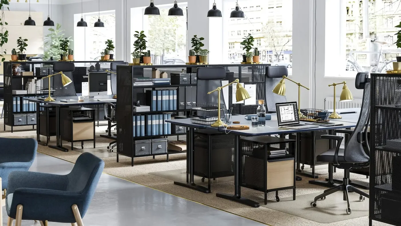 Modern open-plan office with multiple desks, black office chairs, gold desk lamps, green potted plants on high shelves, large windows letting in natural light, and urban street view outside.