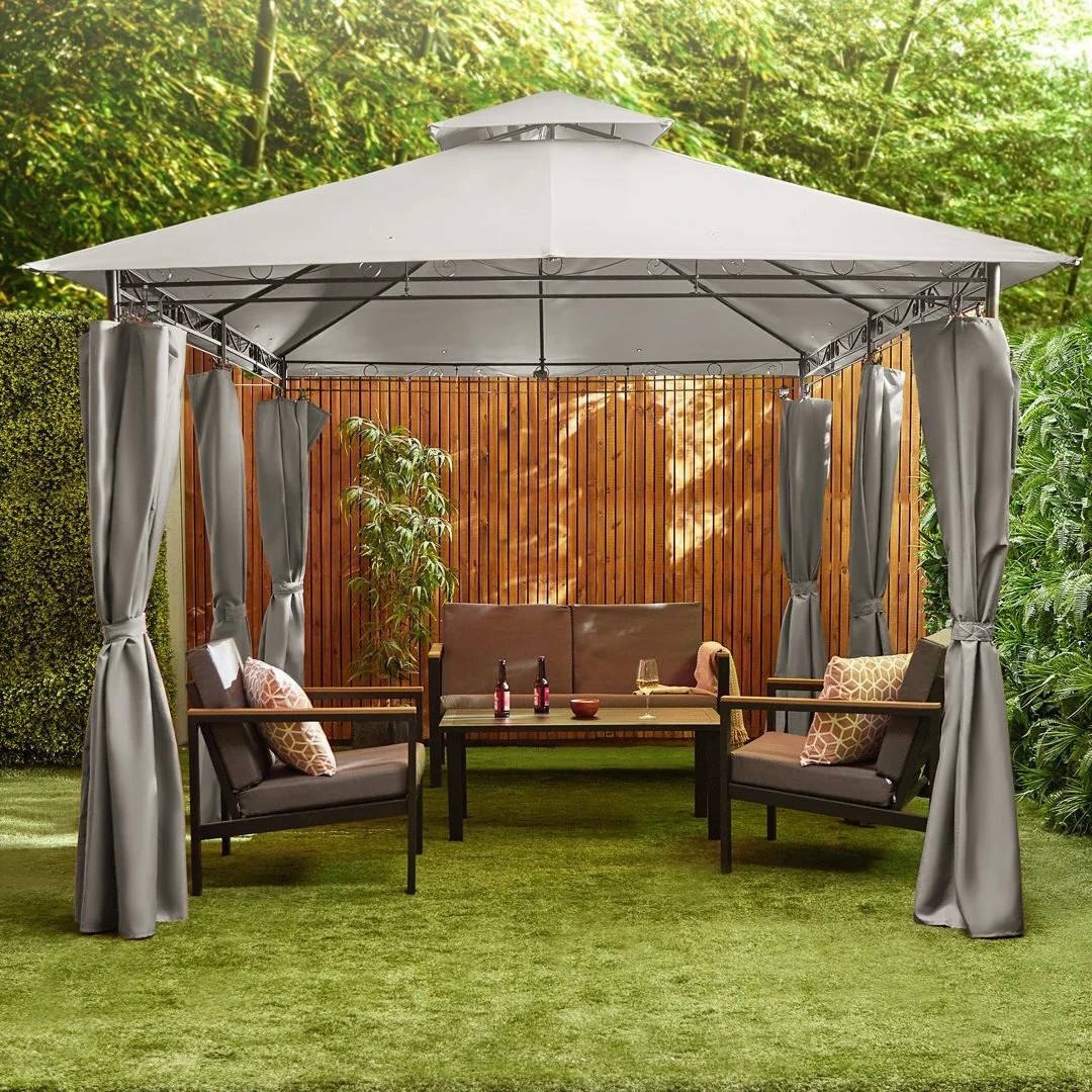 A backyard patio gazebo with curtains, outdoor seating, a small table with drinks, and a wooden fence surrounded by greenery.