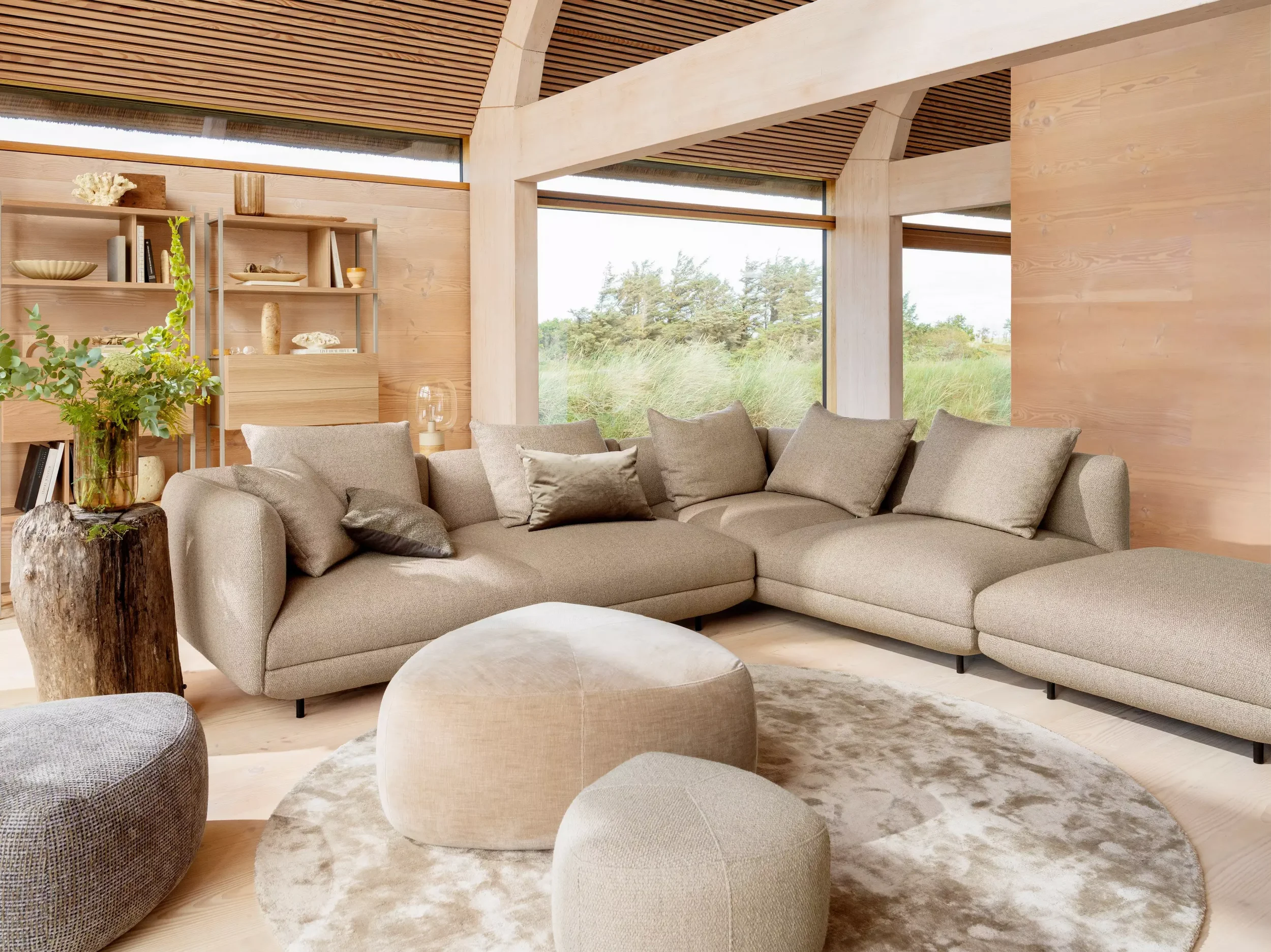 Modern living room with beige sectional sofa, round ottomans, a wooden wall with shelves, large windows showing greenery, and natural wood ceiling and walls.