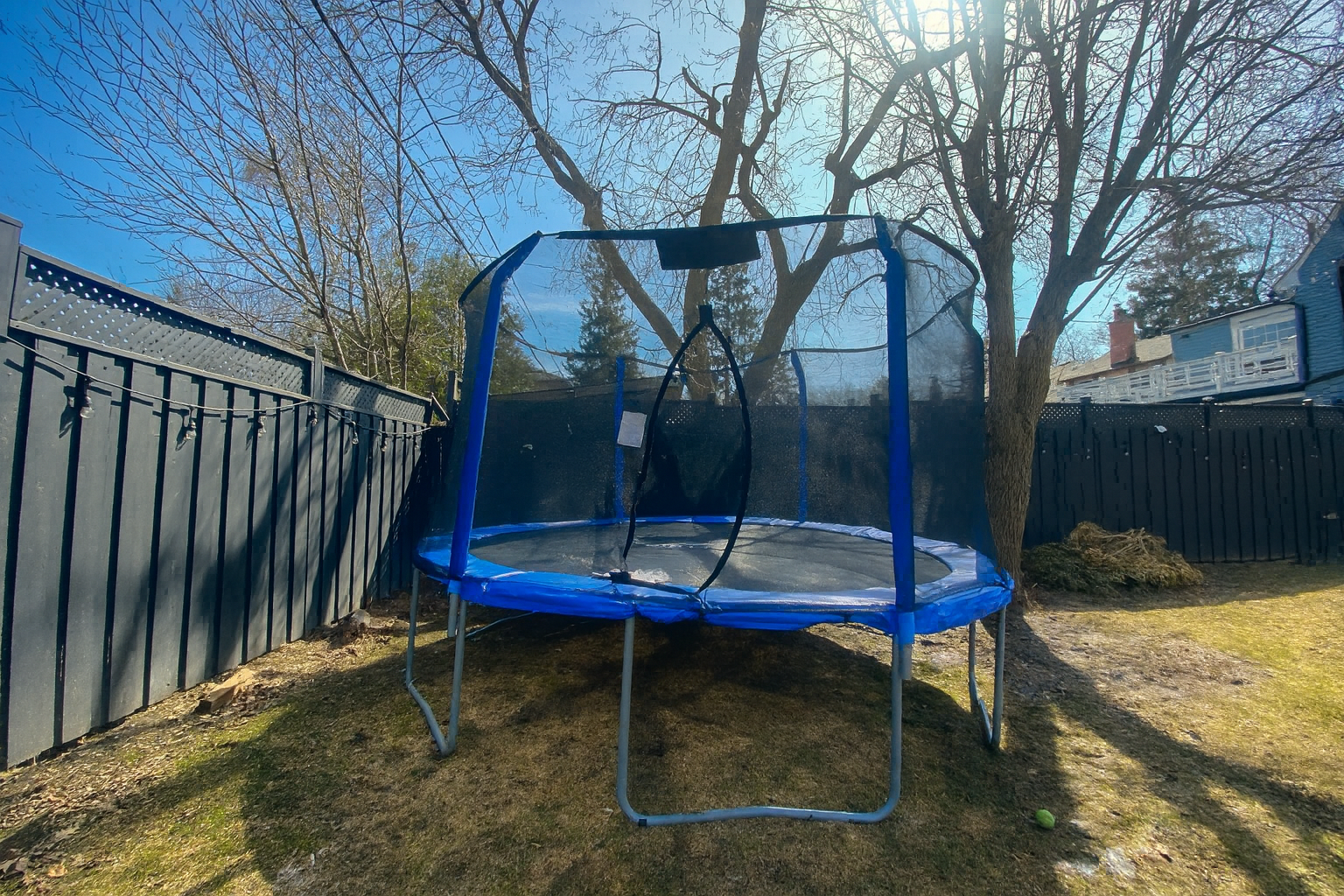 A backyard trampoline with a safety net, surrounded by a black fence and trees, on a sunny day.