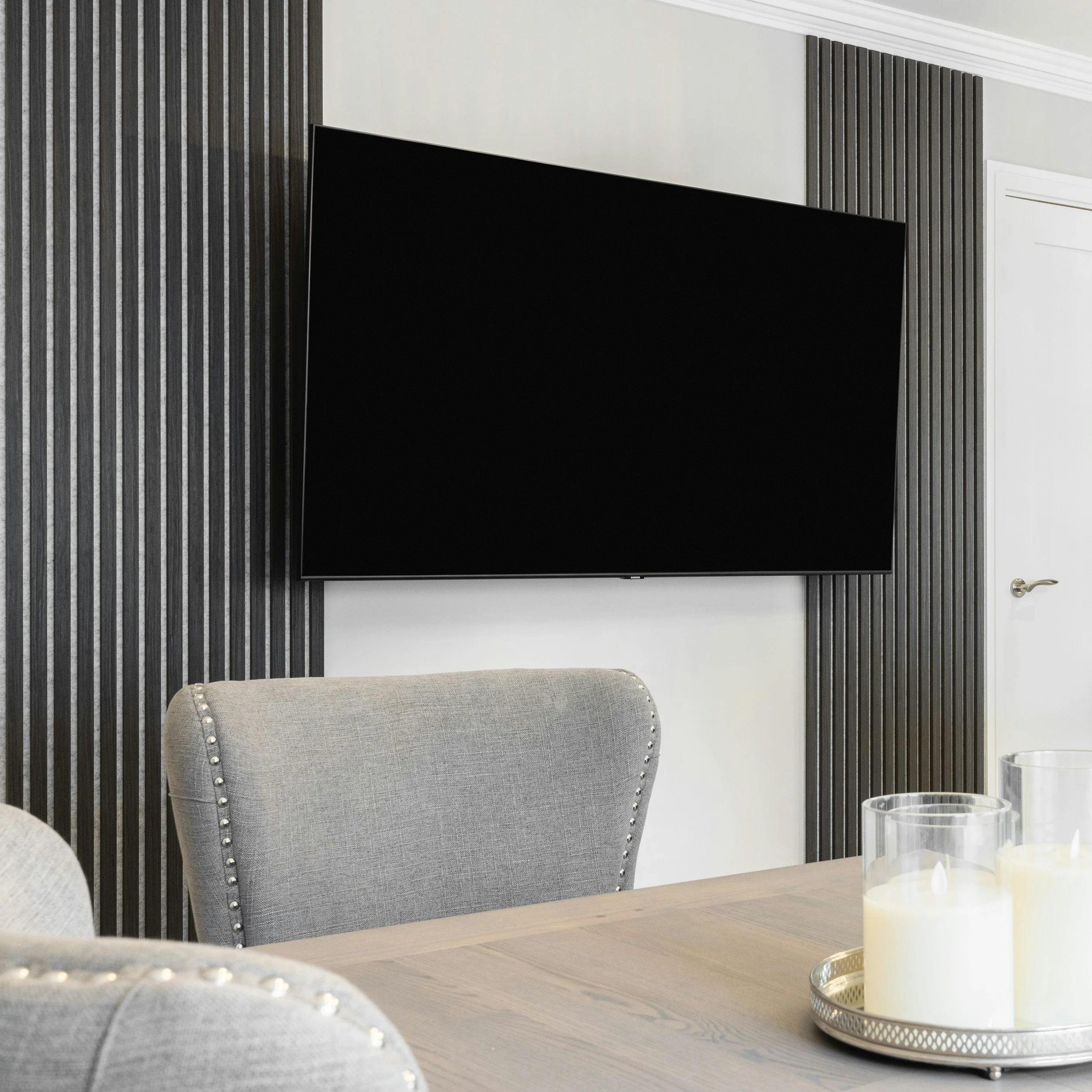 Living room with a large black flat-screen TV mounted on a wall with gray wooden paneling, gray upholstered chair with nailhead trim in the foreground, and candles on a silver tray on a wooden table.