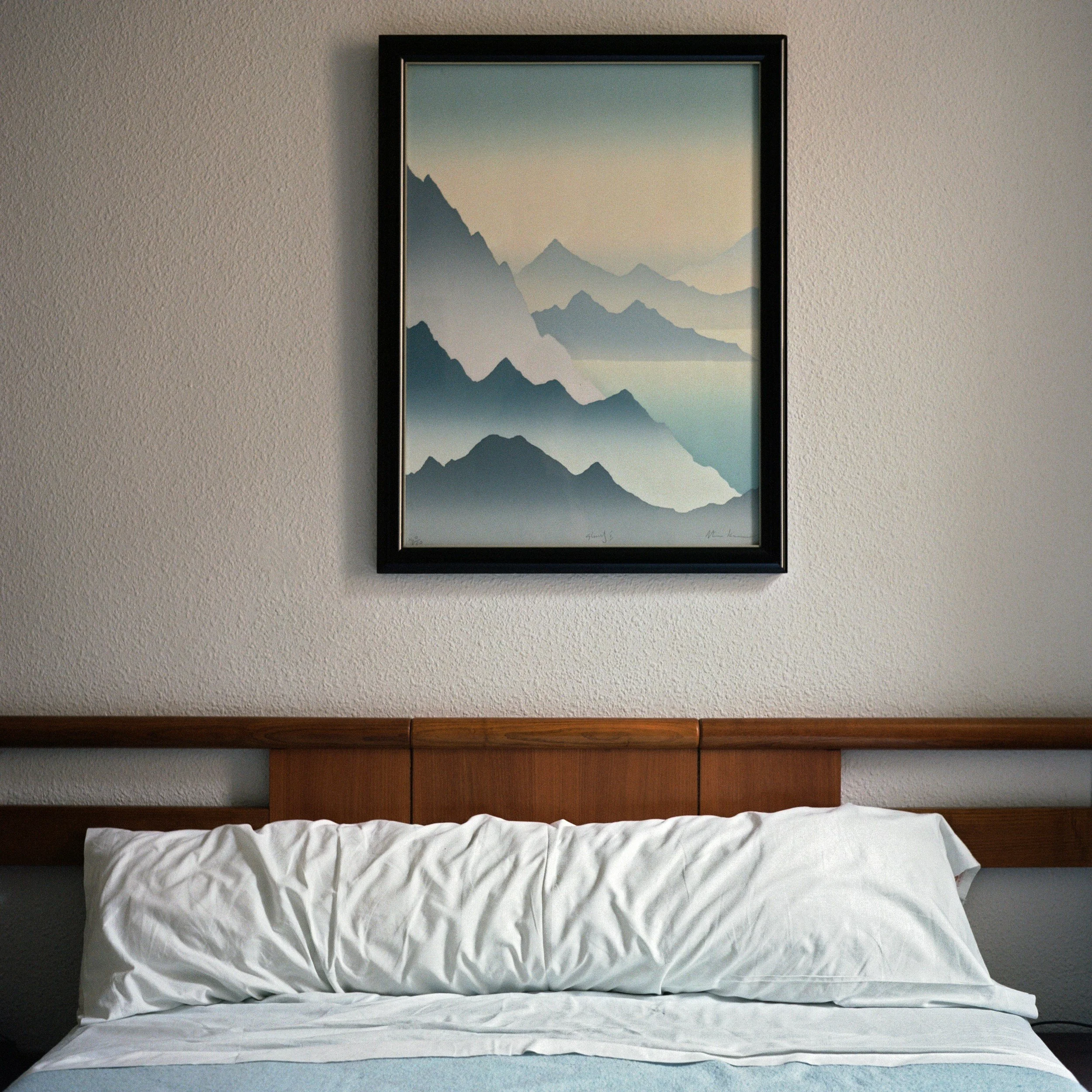 Framed landscape painting of mountain ranges hanging above a bed with a white pillow and blanket, on a textured beige wall.