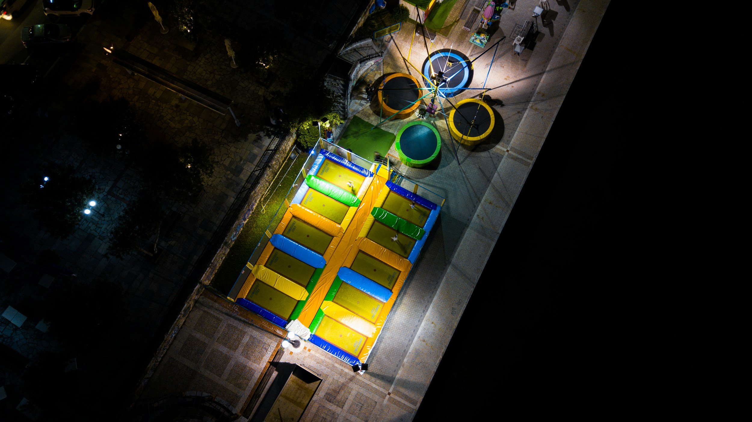 An aerial view of an illuminated urban playground at night showing a large inflatable obstacle course, several trampoline stations, and tunnel structures, with surrounding streets and trees.