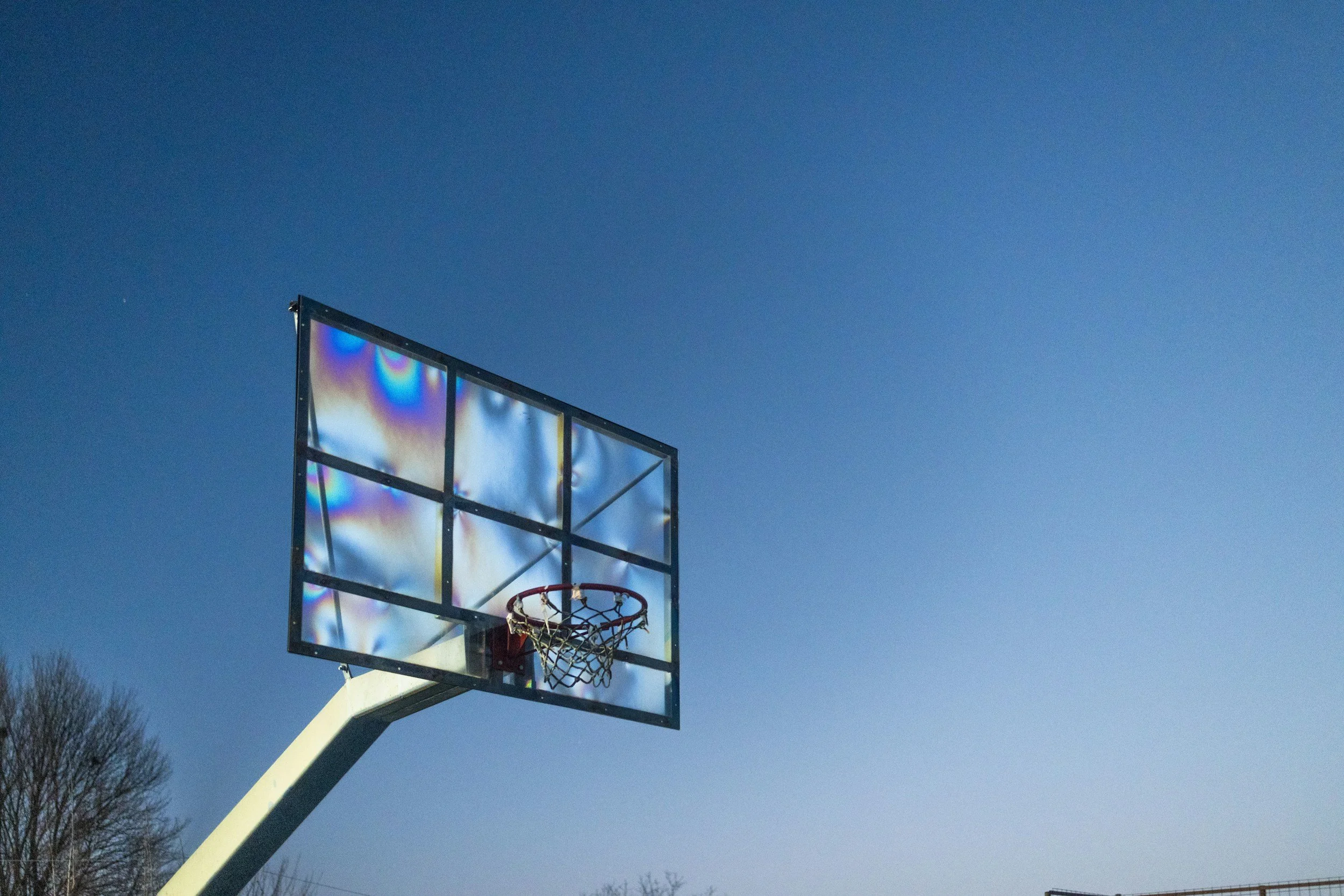 An outdoor basketball hoop with a backboard reflecting sunlight, set against a clear blue sky and some trees in the background.