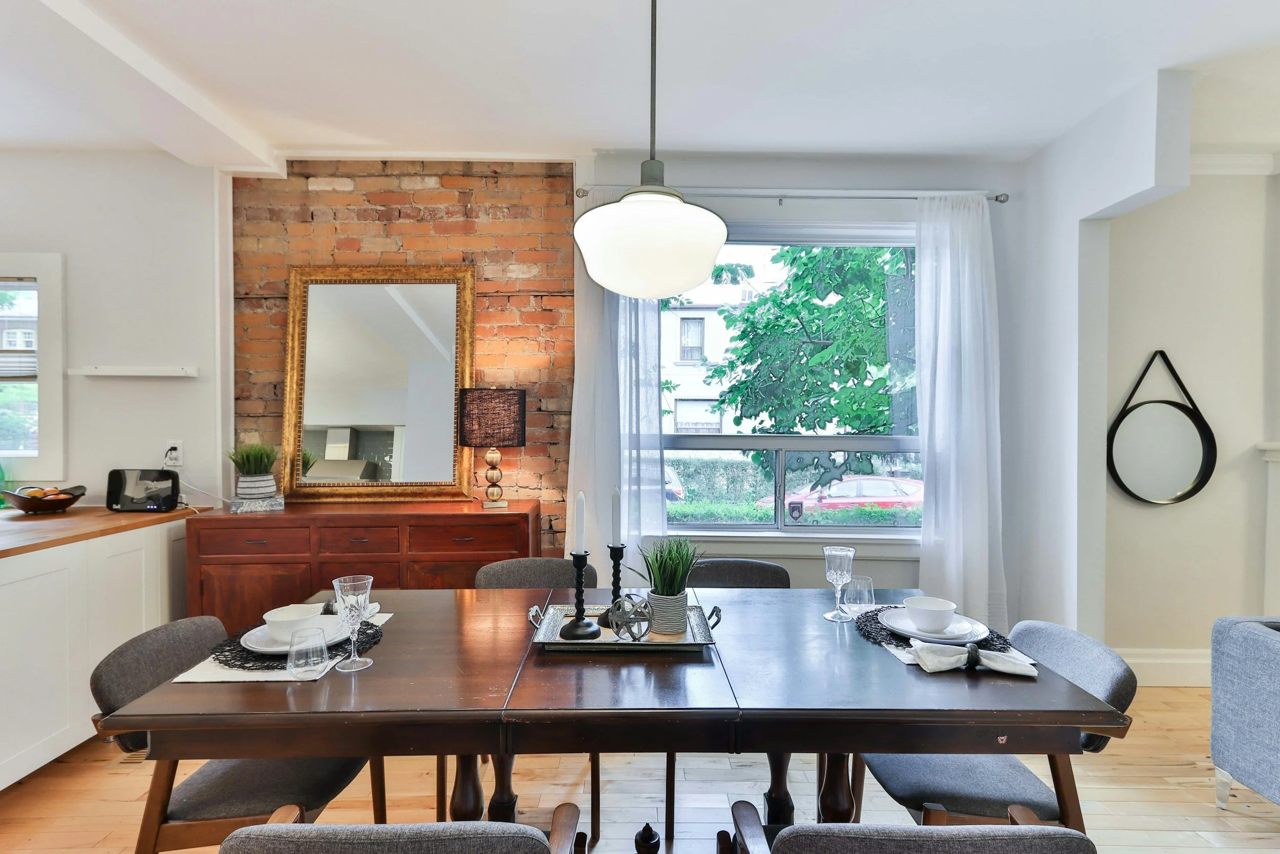 Dining room with wooden table set with plates, glasses, and black placemats, surrounded by gray chairs, featuring a large window with white curtains, brick accent wall with mirror, sideboard with lamp, and decor items, with hardwood floors and white walls.