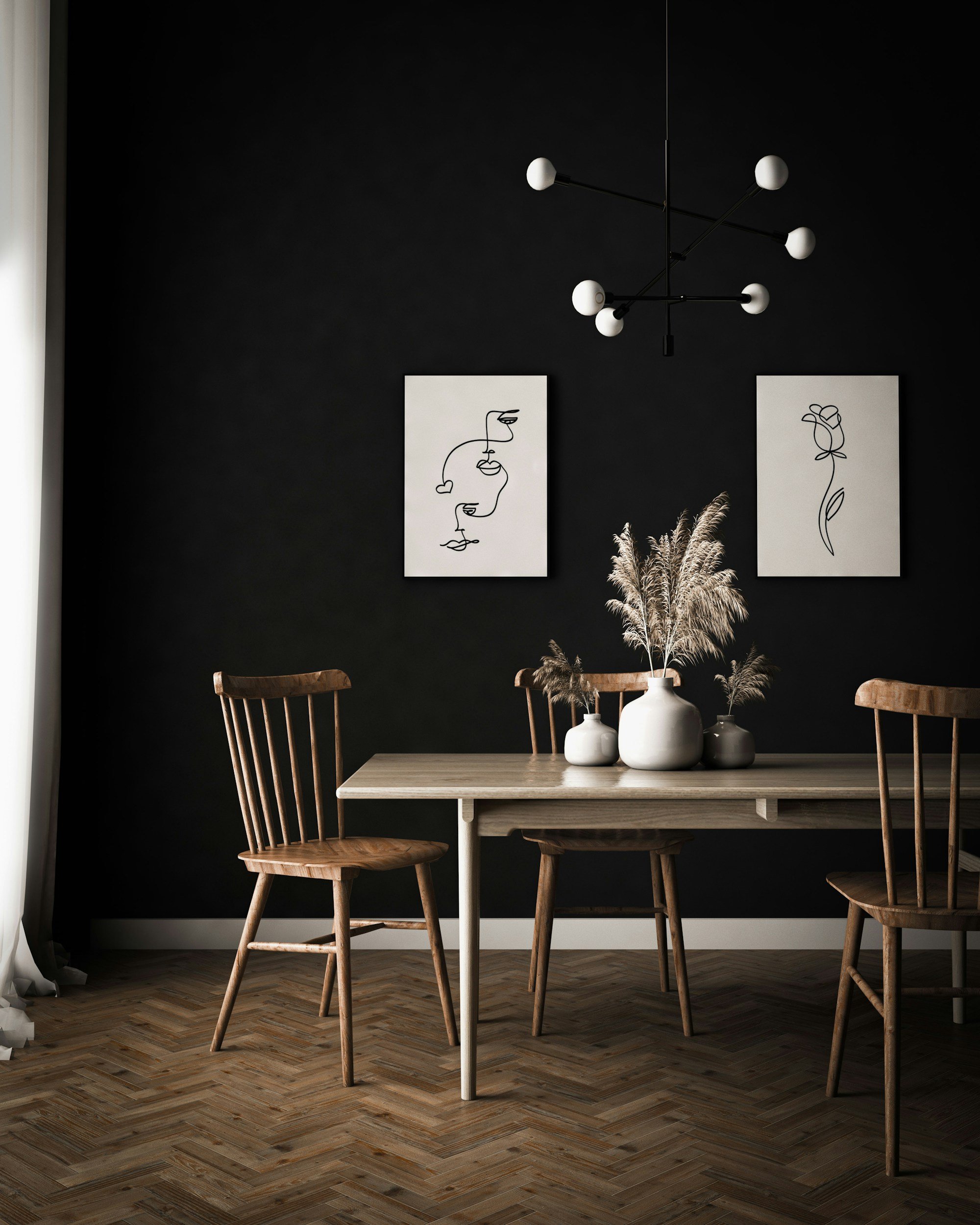 A dining room with a light wooden table and three wooden chairs. The table has three white vases with pampas grass. The background has a dark wall with two minimalist line art drawings of faces and a flower, and a modern chandelier with multiple white globe lights hangs from the ceiling.