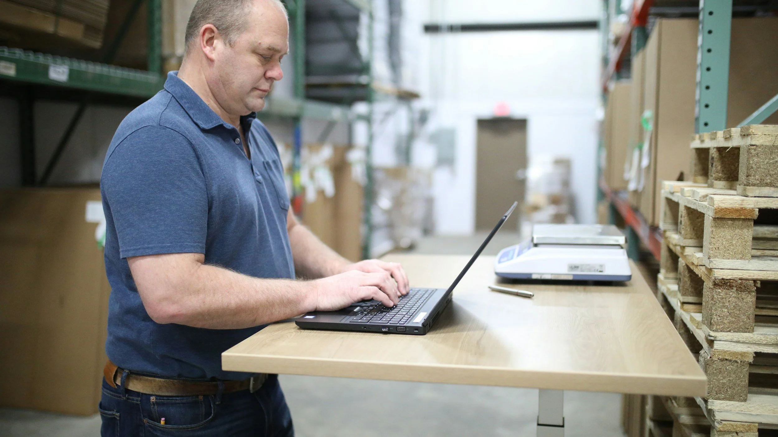 A man in a blue polo shirt working on a laptop at a wooden workbench in a warehouse or storage area with shelves of boxes and wooden pallets.