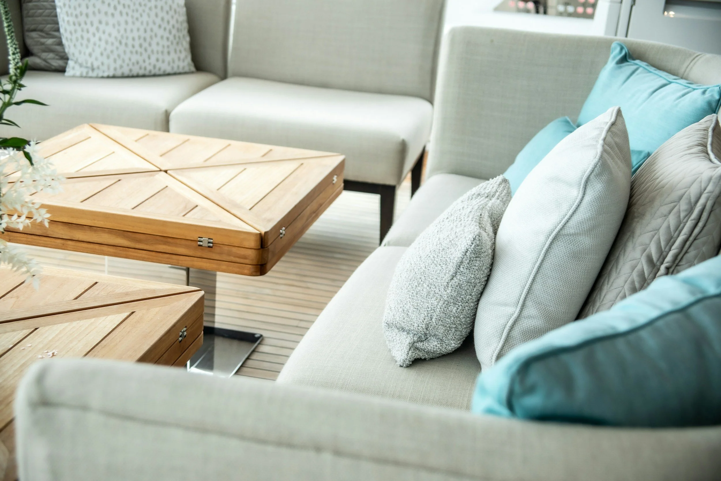 Living room with beige sofas and wooden coffee tables, decorated with various cushions in white, light gray, and turquoise, and a potted plant visible on the left.
