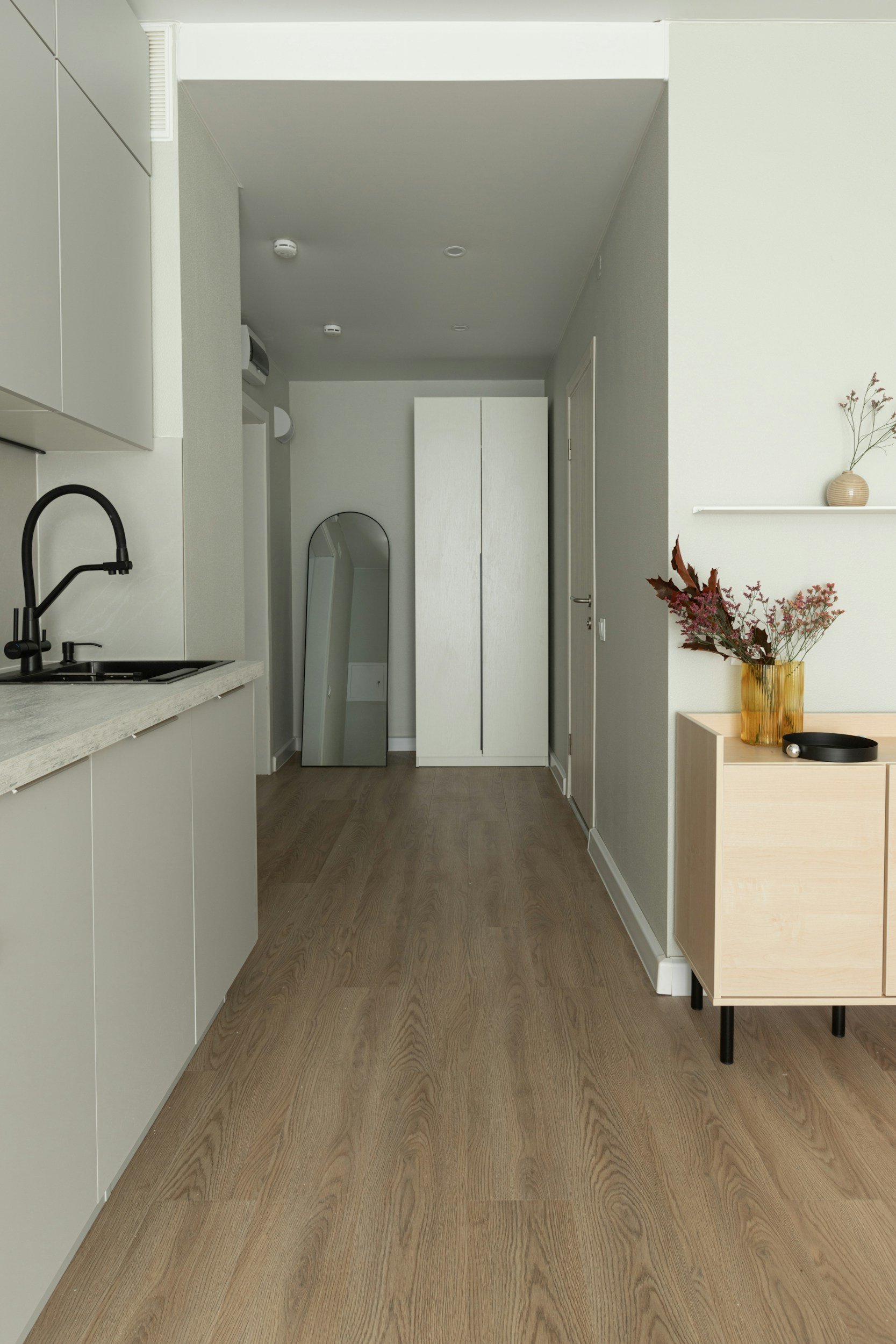 Minimalist kitchen and hallway with wooden flooring, white walls, black faucet, tall white cabinet, leaning oval mirror, beige side table with floral arrangement, and a door in the background.
