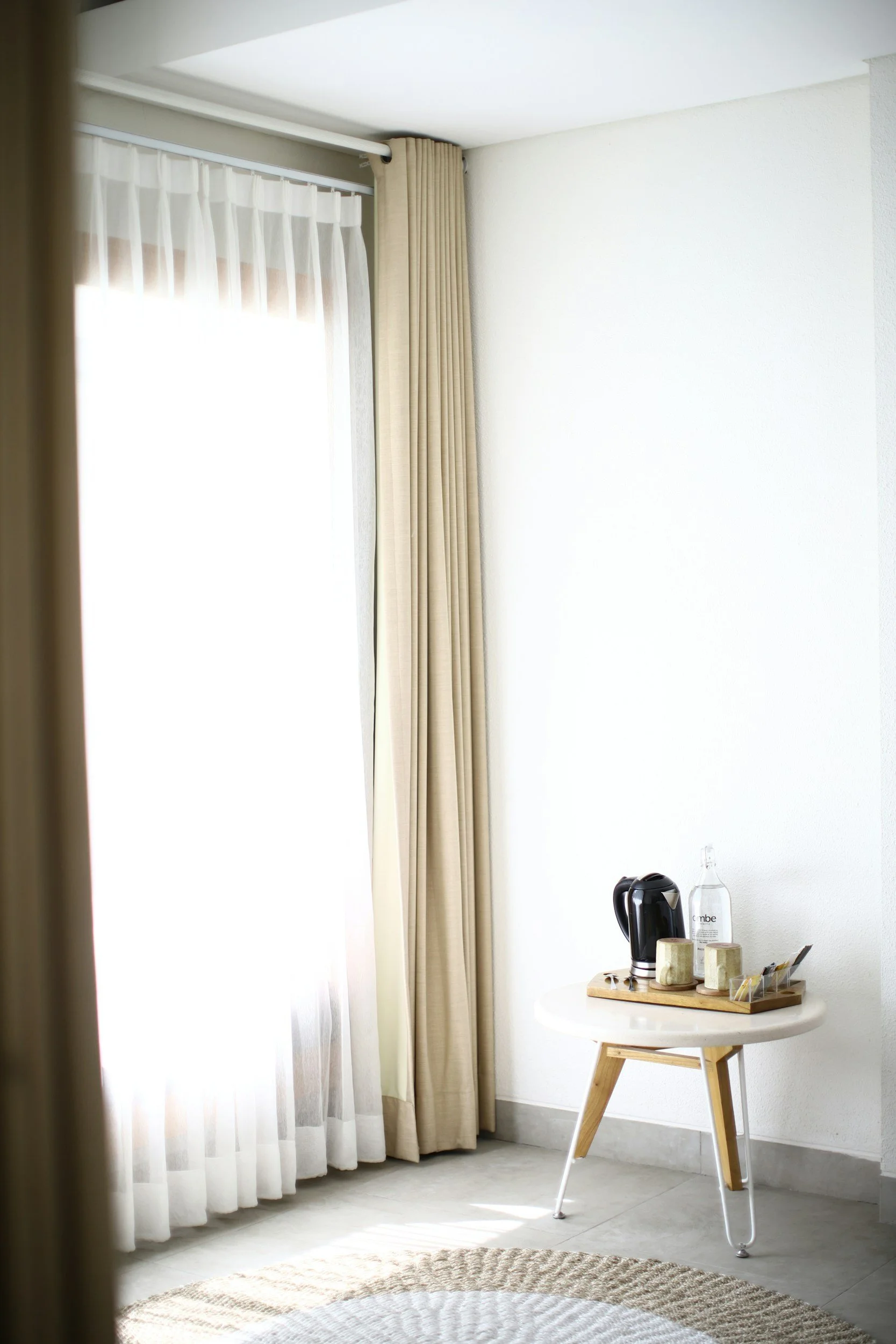 Sunlit room with beige curtains, a small round white table holding a black electric kettle, glass bottle of water, and cups, near a white wall and beige rug.