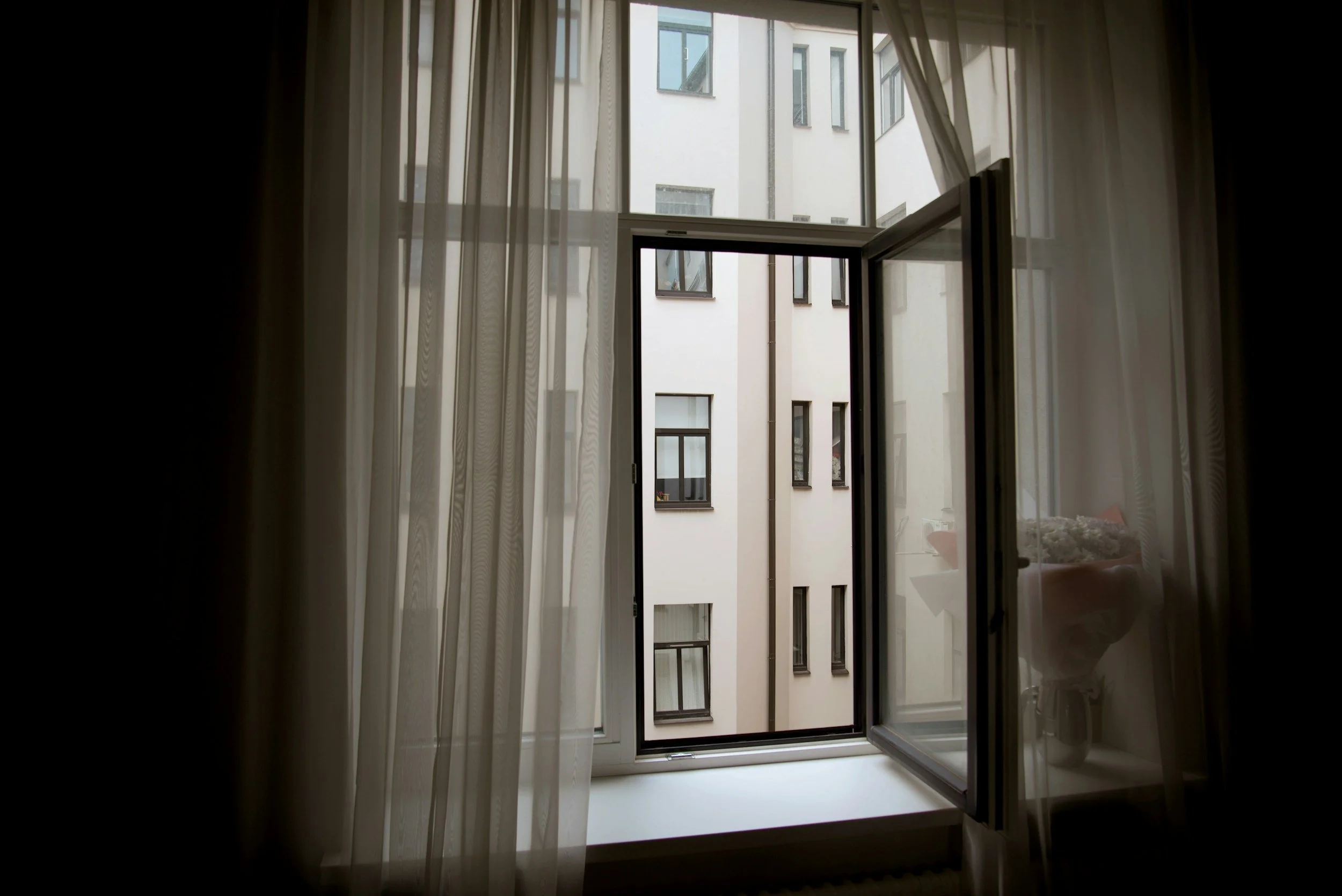 Open window with sheer curtains revealing a view of an apartment building with multiple windows.
