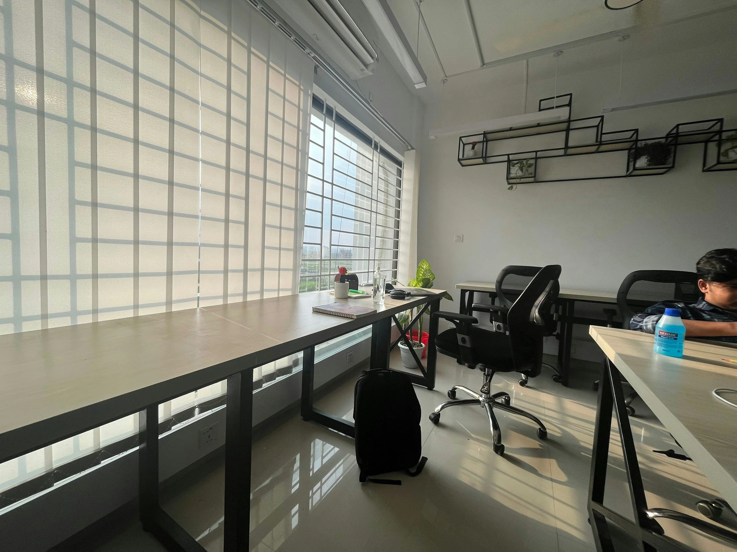 An office room with large windows letting in sunlight, with desks, a backpack, office chairs, and some plants.