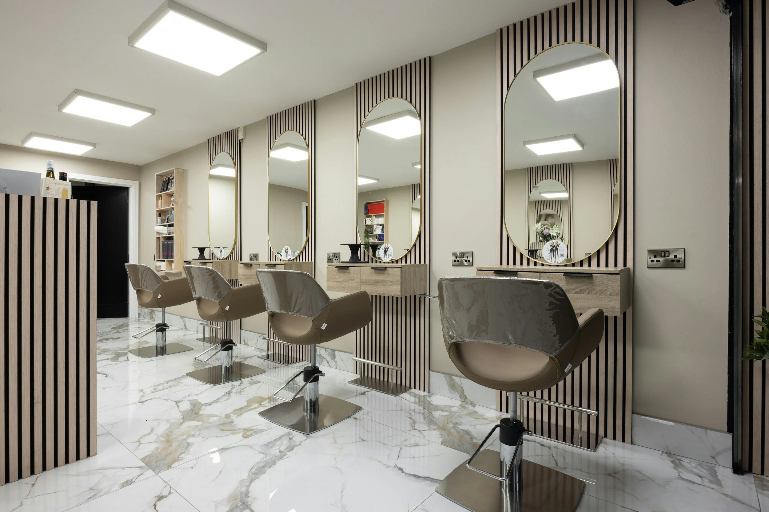 Modern salon with three styling chairs in front of tall, oval mirrors mounted on wooden slat wall panels, with a marble floor and overhead lighting.