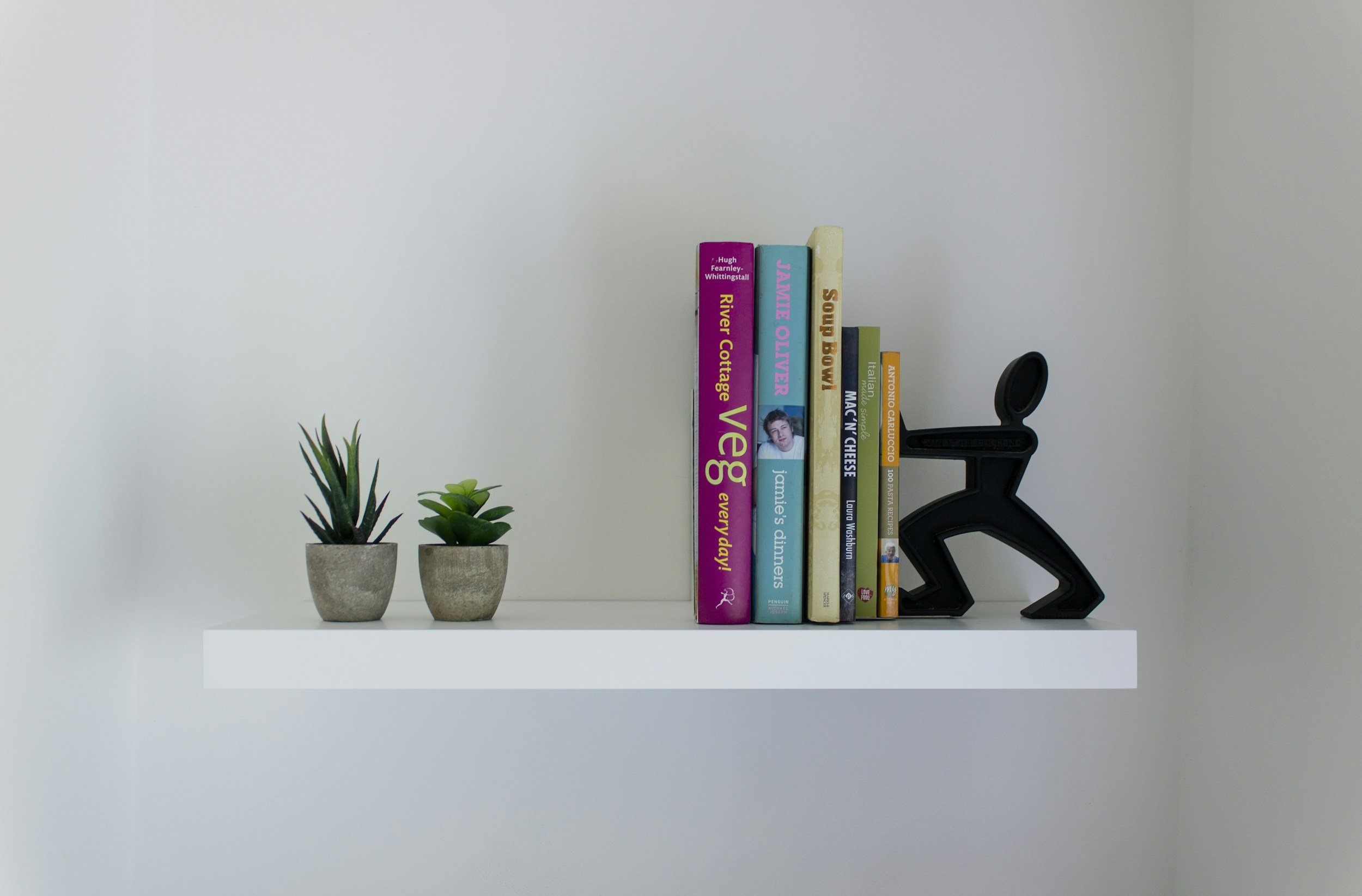 White floating shelf holding two small potted succulents on the left, a row of colorful books in the middle, and a black abstract sculpture of a person on the right against a plain white wall.