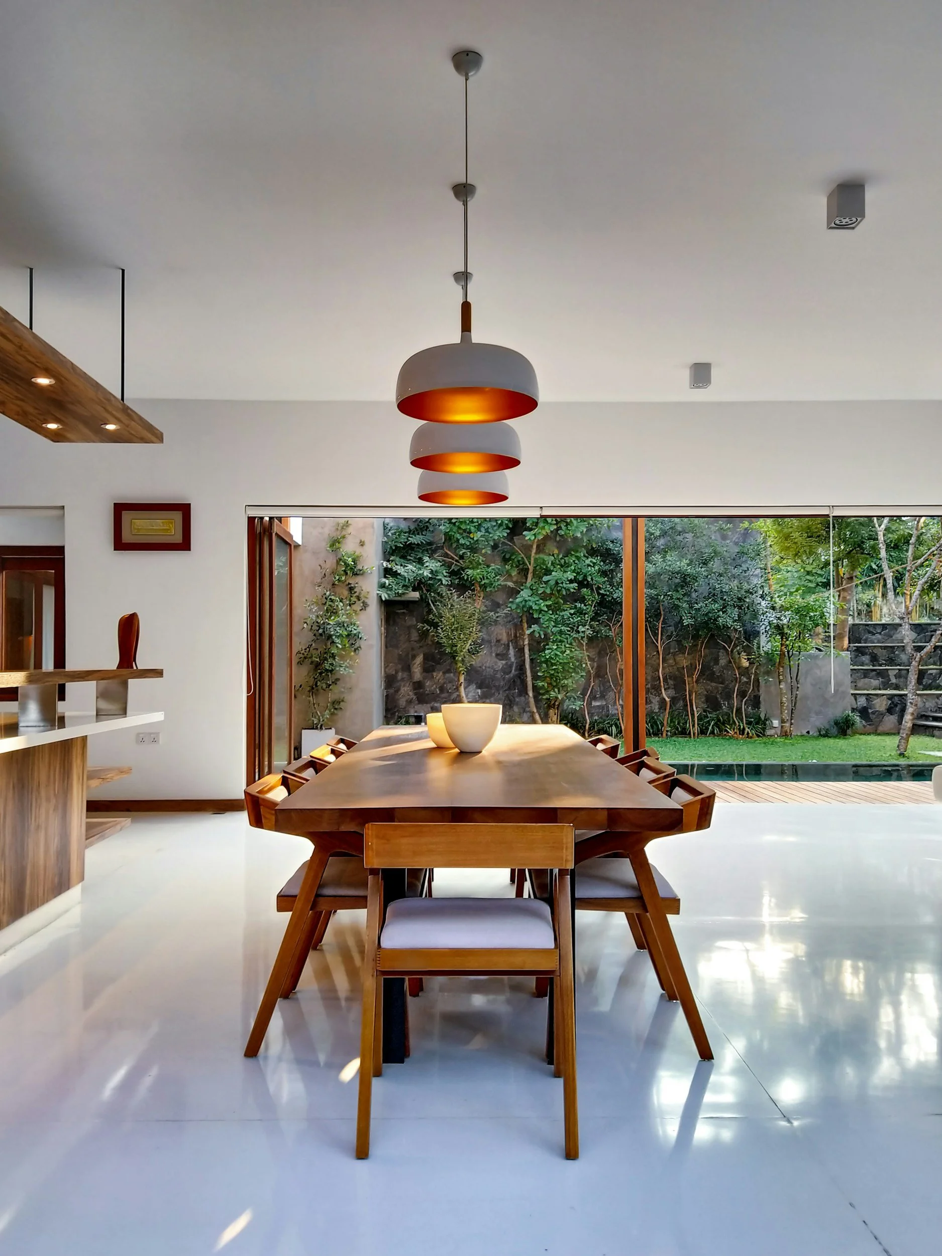 Modern dining room with a large wooden table, white chairs, and a three-tier hanging pendant light fixture above. Large glass sliding door opens to a lush outdoor garden.