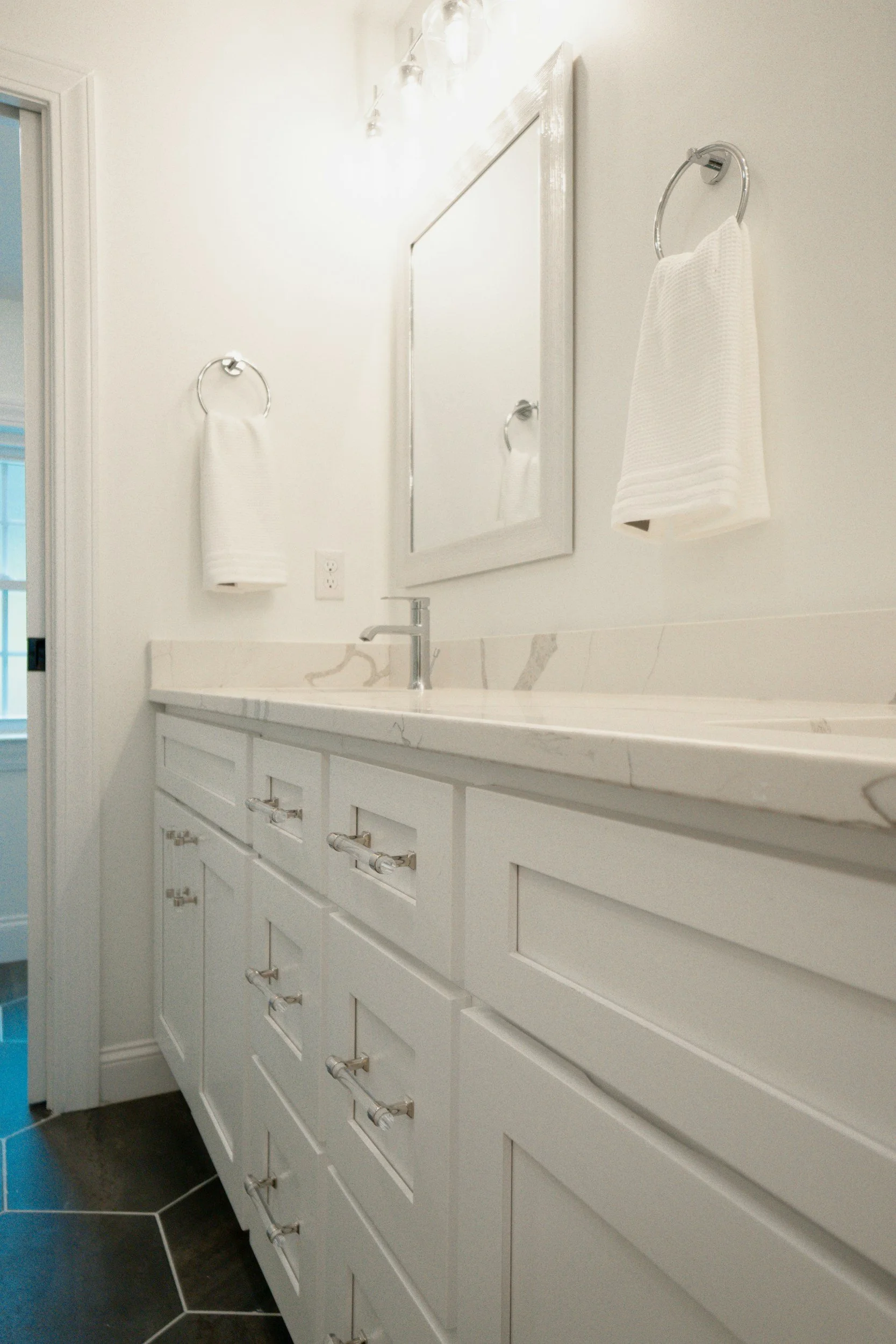 White bathroom vanity with multiple drawers and cabinets, a marble countertop, a silver faucet, a large mirror with white frame, and two white hand towels on silver ring holders mounted on the wall.