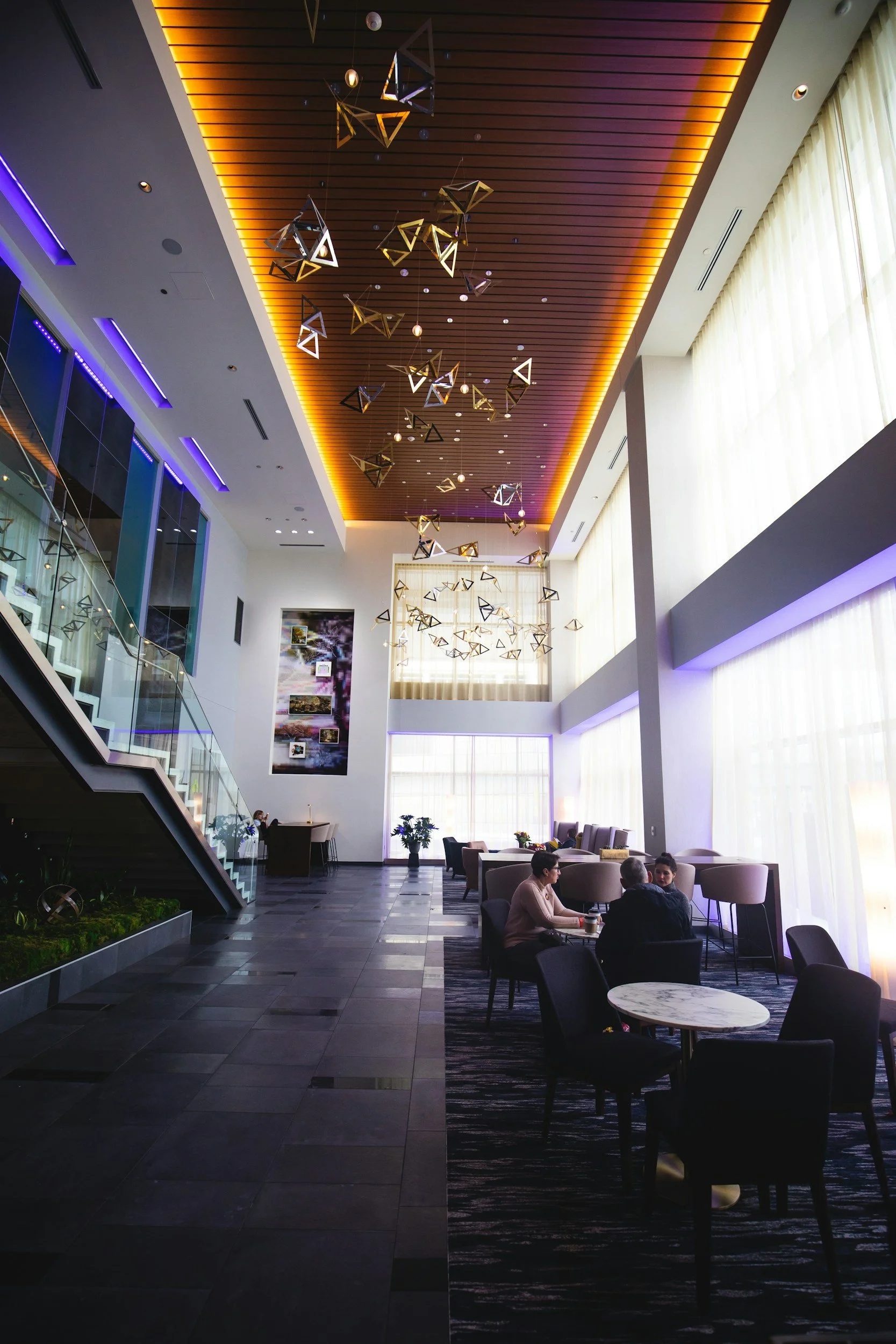 Modern hotel lobby with high ceilings, hanging geometric light fixtures, large windows with sheer curtains, and a seating area with tables and chairs, where a few people are sitting and talking.