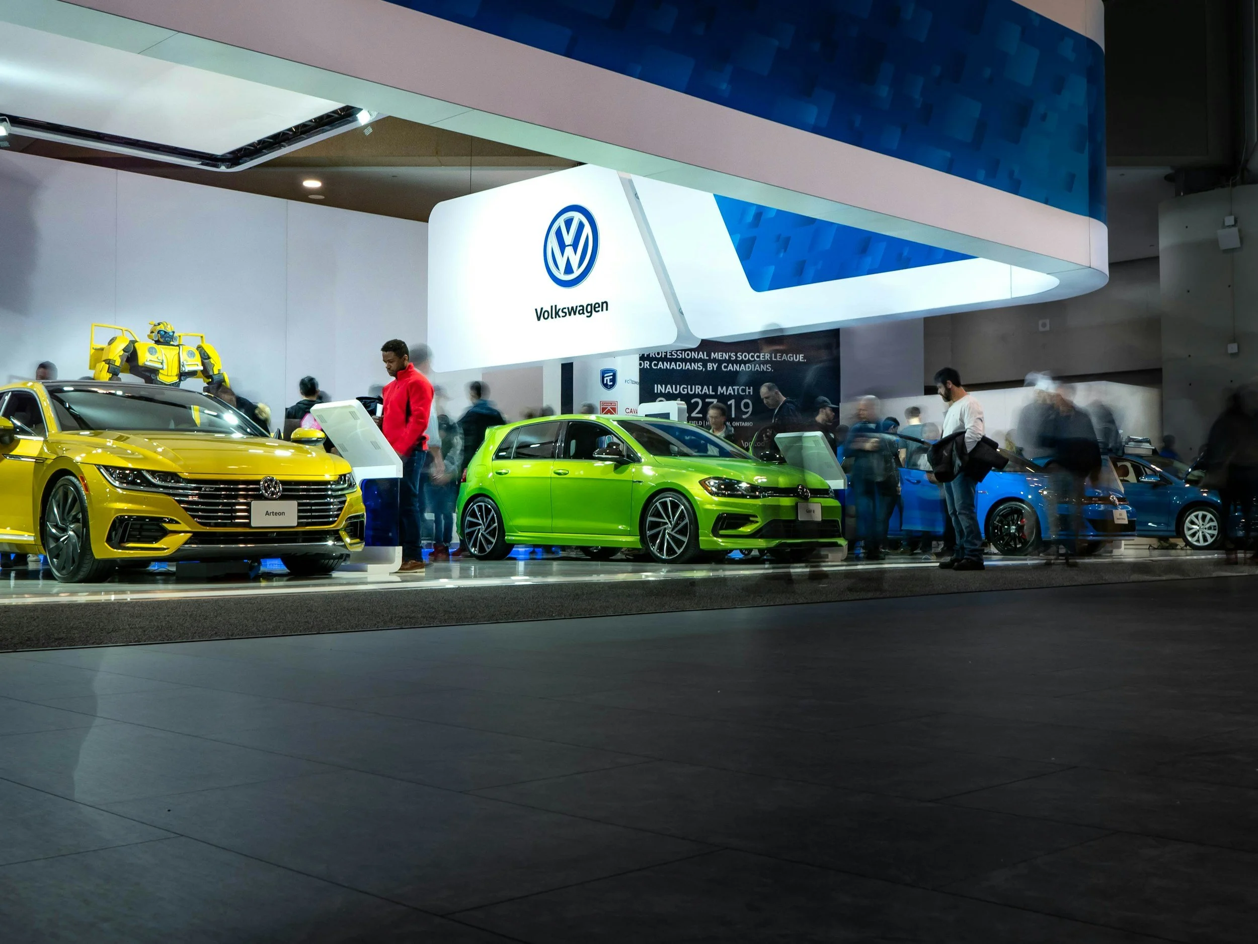 A display booth at an auto show featuring Volkswagen cars, including a yellow sedan, a green hatchback, and a blue sedan, with people walking around and inspecting the vehicles.