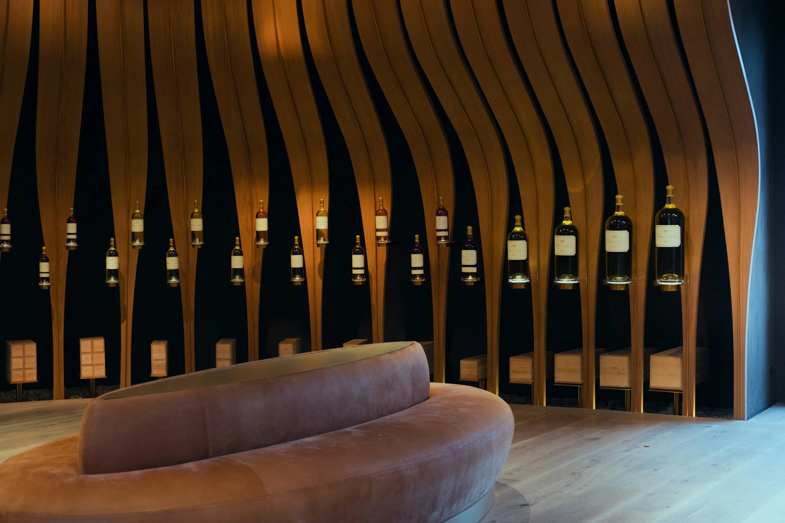 A modern wine cellar with wooden wall panels, wine bottles mounted on the wall, and a pink curved sofa in the foreground.