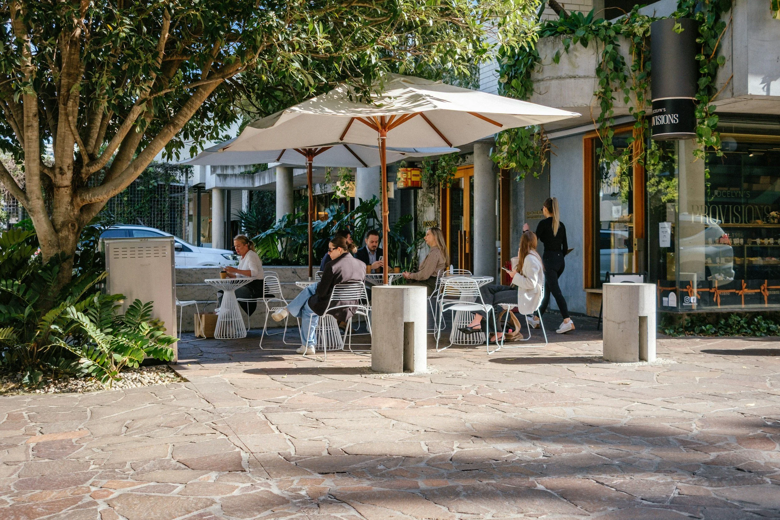 People sitting at outdoor tables under large umbrellas at a cafe on a sunny day, surrounded by trees and greenery outside a storefront.